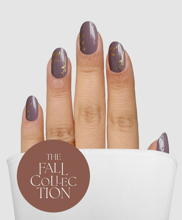 A hand with short nails featuring the Sassy Saints - Brings The Salon Home Gilded Mauve Gel Wrap, adorned with delicate gold leaf accents, emerges from a white pocket. A brown circle displays the text: The Fall Collection.