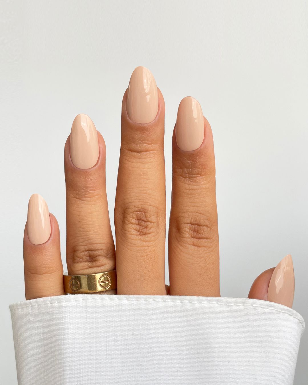 A hand with Sassy Saints - Brings The Salon Home Naked Ambition Gel Wrap nails, glossy and nude-colored, is shown against a plain white background. A gold ring adorns one finger and a white shirt sleeve partially covers the hand.