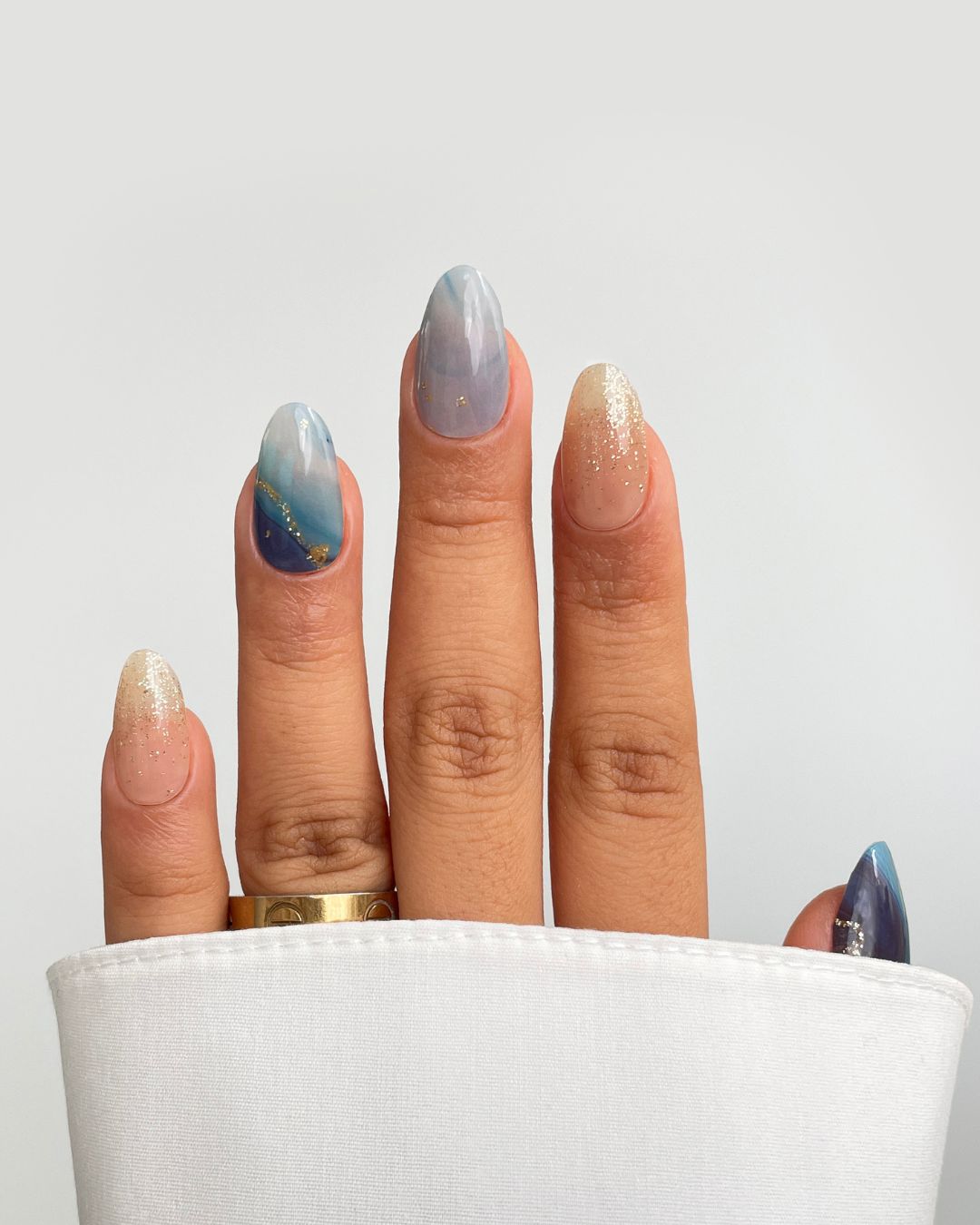 A hand with Sassy Saints - Brings The Salon Home's Ocean Dream Gel Wrap—almond-shaped nails with a blue and white gradient, gold accents, and glitter. A gold ring is worn on the ring finger, partially covered by a white sleeve.