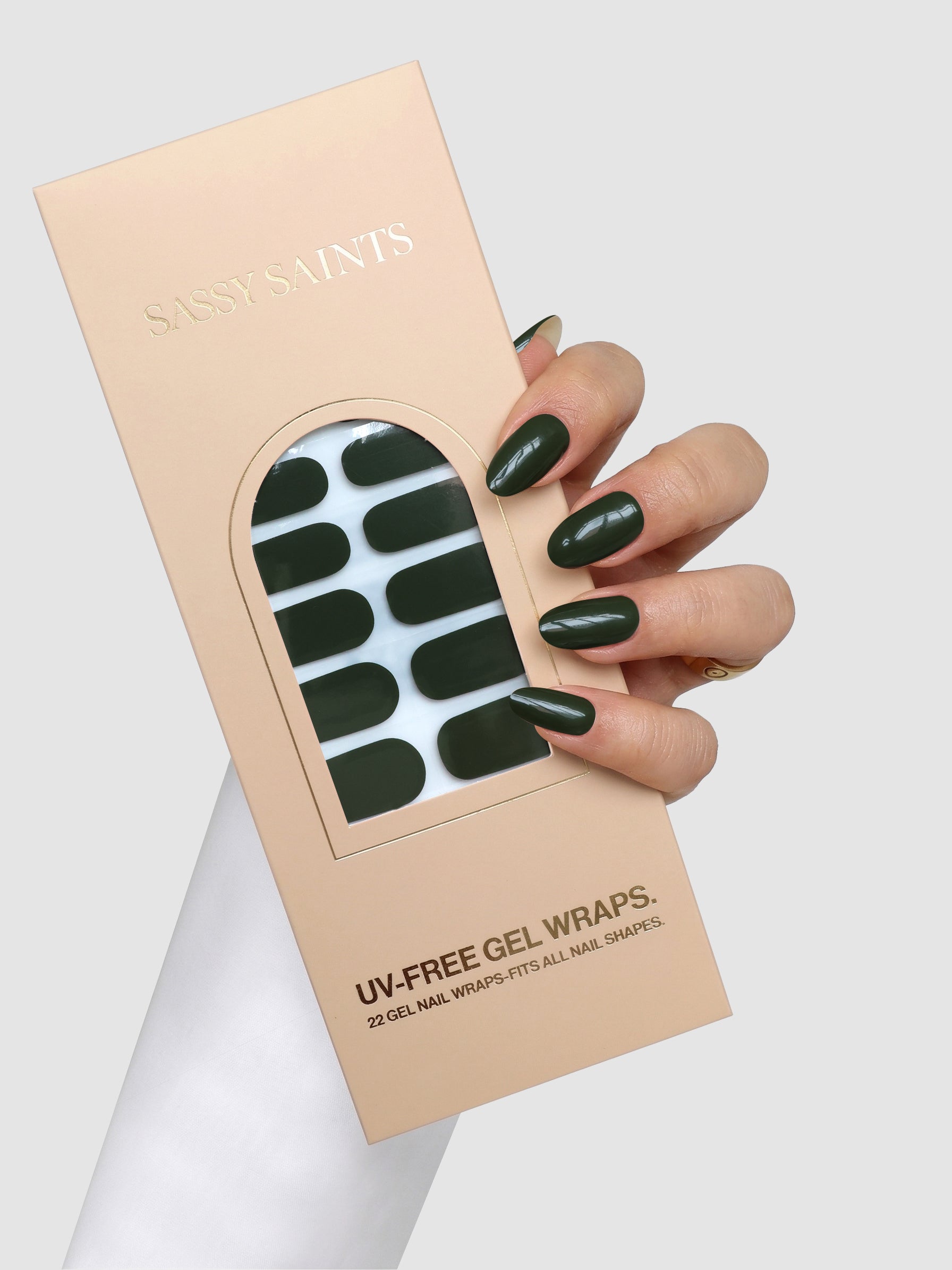 A hand with dark green nails holds a Sassy Saints - Brings The Salon Home Olive Instinct Gel Wrap box containing black oval UV-free gel nail wraps against a light gray background.
