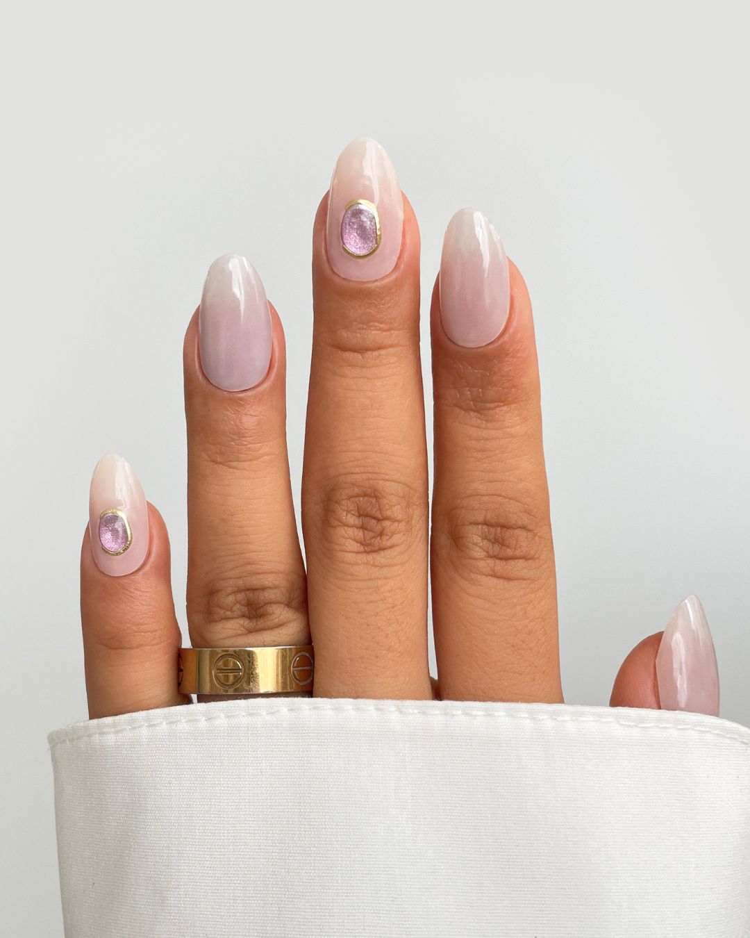 A hand with Sassy Saints' Opal Gem Gel Wrap nails features glossy, almond-shaped nude-pink wraps and light purple gem accents on the ring and thumb, paired with a gold ring and a white long-sleeve top.