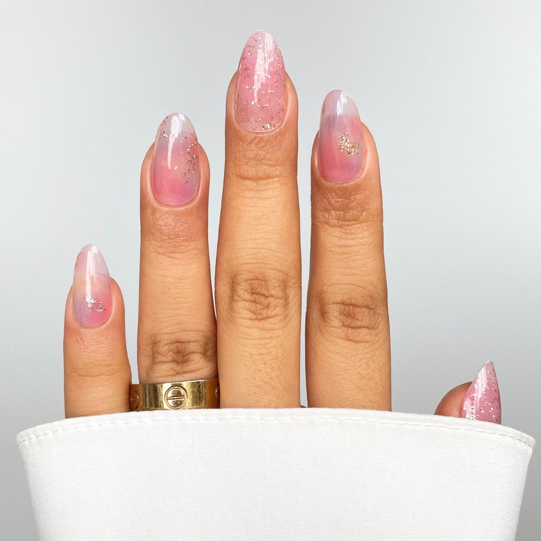 A hand with long, almond-shaped nails features Sassy Saints - Brings The Salon Home’s Pastel Haze Gel Wrap in sheer pink glitter. A gold ring adorns the middle finger, and a white shirt sleeve partially covers the hand.
