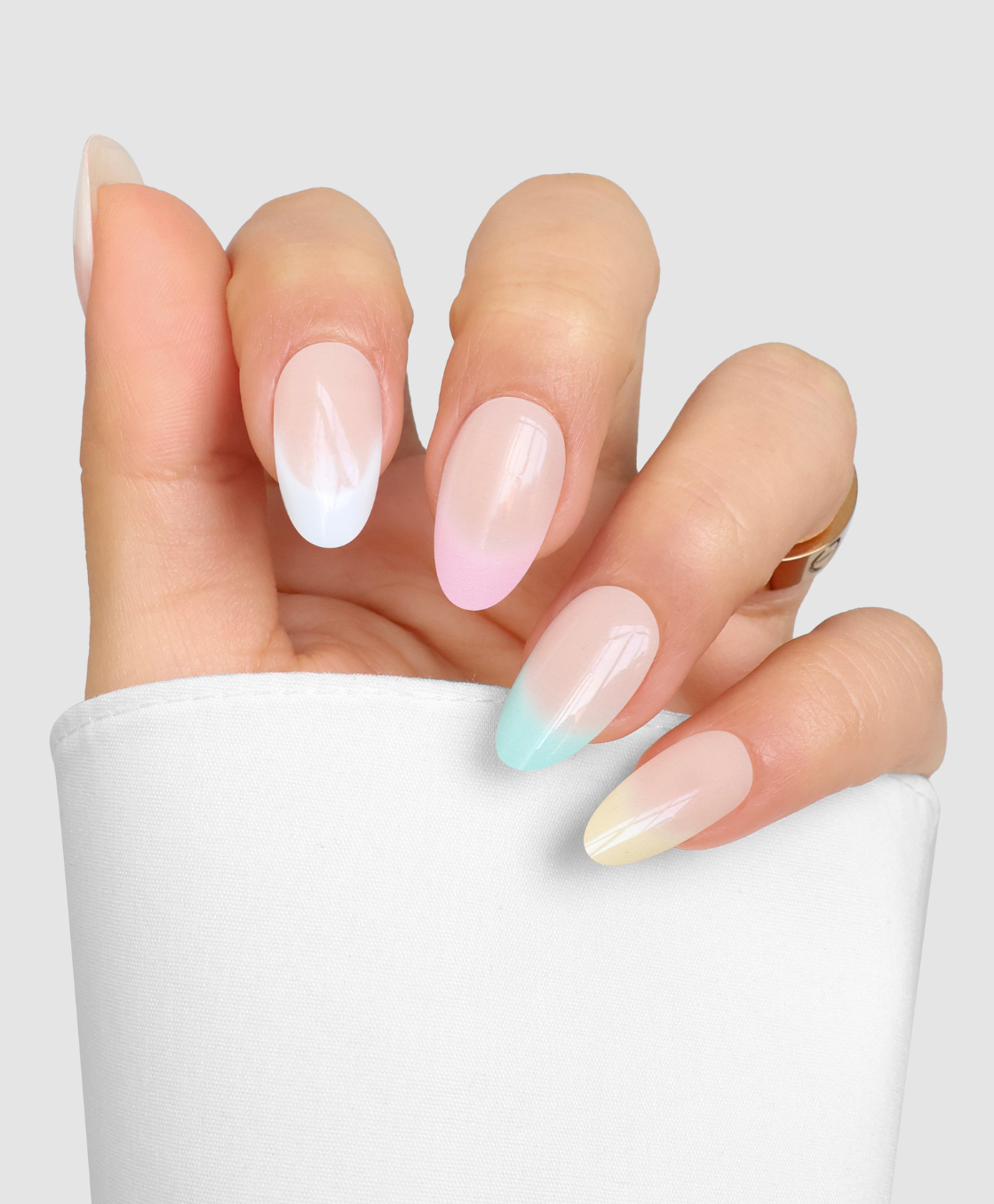 A hand with long, almond-shaped nails features Sassy Saints' Pastel Parade—ombre blue, pink, yellow, and mint green—against a light gray background. The person wears a white sleeve and a gold ring.