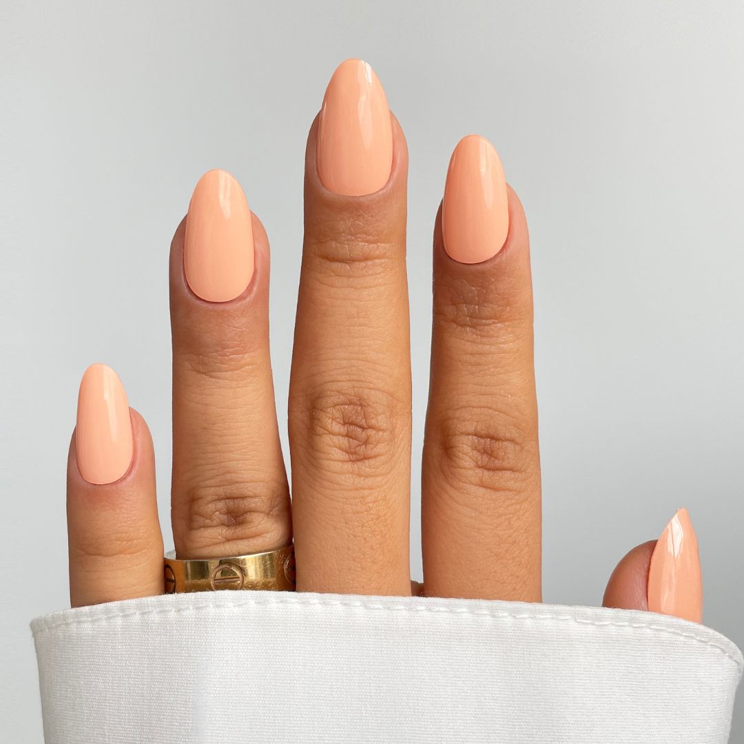 A hand shows off Sassy Saints' Peach Glow Gel Wrap nails in a glossy almond shape, partially covered by a white sleeve. A gold ring adorns the middle finger against a plain light gray background.