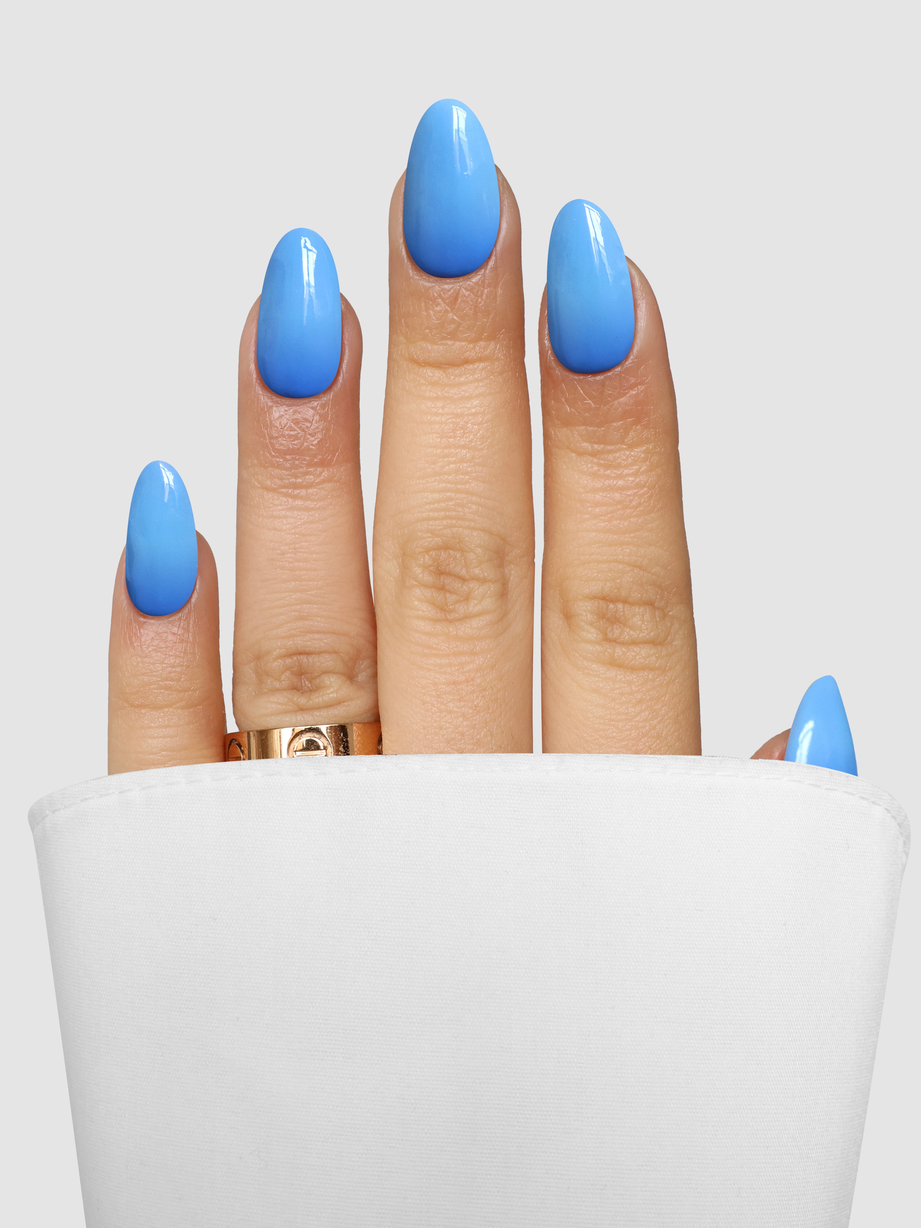 A hand with glossy, oval-shaped nails in Sassy Saints’ Periwinkle Pretty Dipping Powder peeks from a white cloth, gold ring on the middle finger, set against a plain gray background.