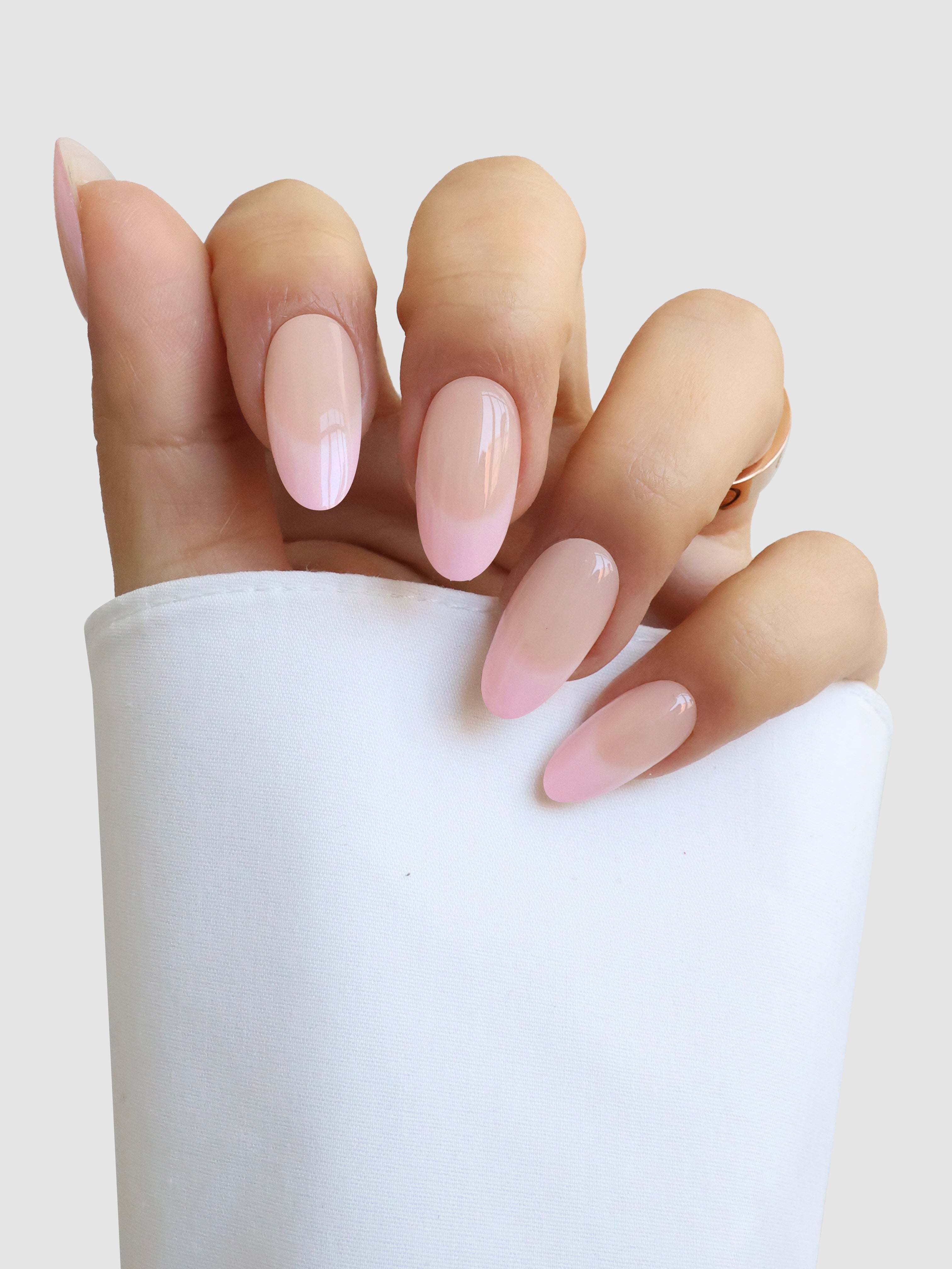 A hand with long, almond-shaped nails painted in glossy light pink Perle de Paris Gel Wrap by Sassy Saints - Brings The Salon Home rests against white fabric, fingers gently curved.