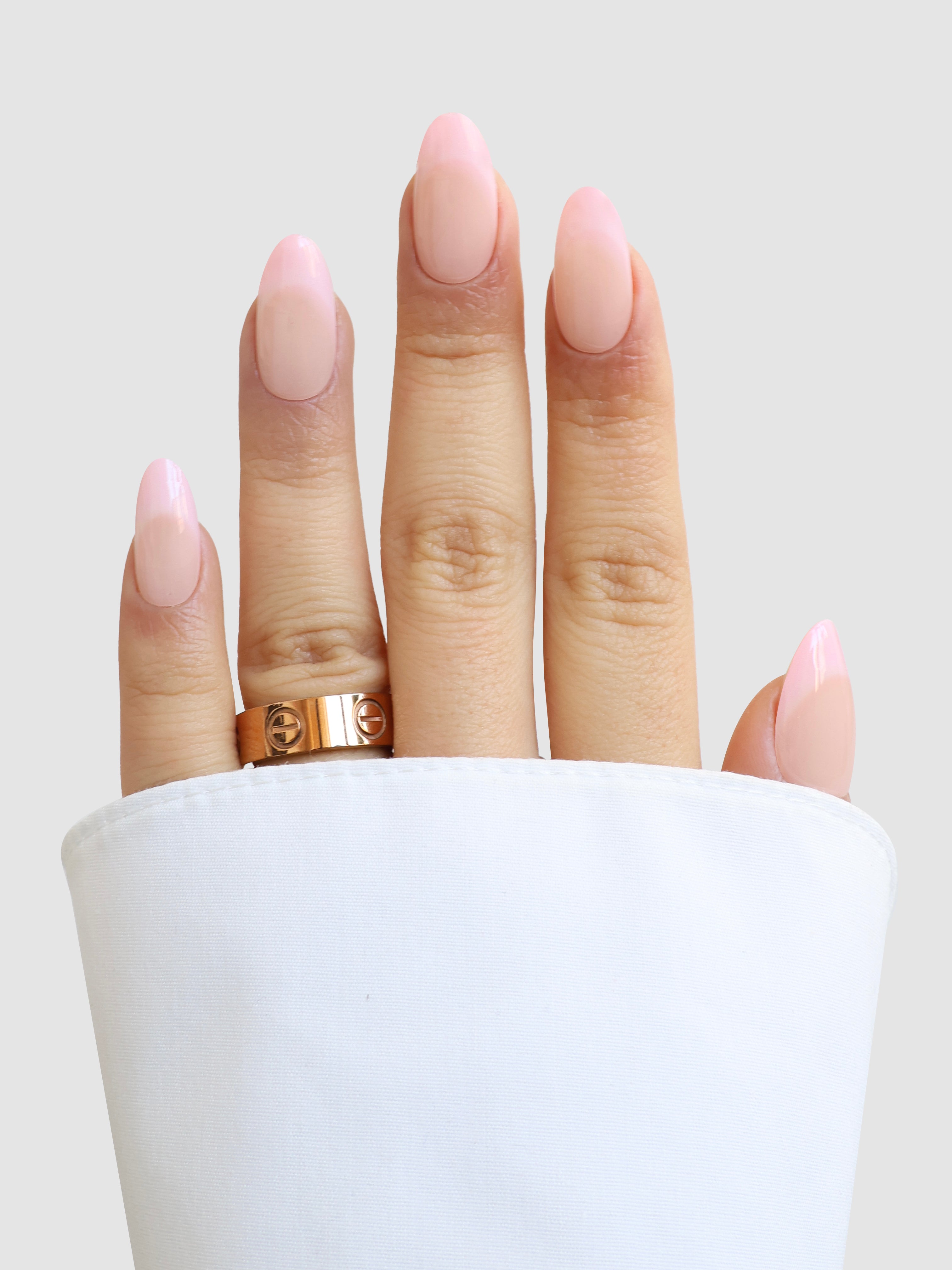 A hand with long, almond-shaped nails in pale pink Perle de Paris Gel Wrap by Sassy Saints - Brings The Salon Home wears a gold ring, partially covered by a white shirt sleeve, set against a light gray background.