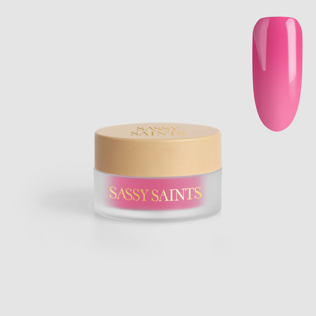 A small round jar labeled “Sassy Saints - Brings The Salon Home” with a beige lid, containing Pink Parfait Dipping Powder, is shown beside a glossy pink swatch on a nail-shaped sample.