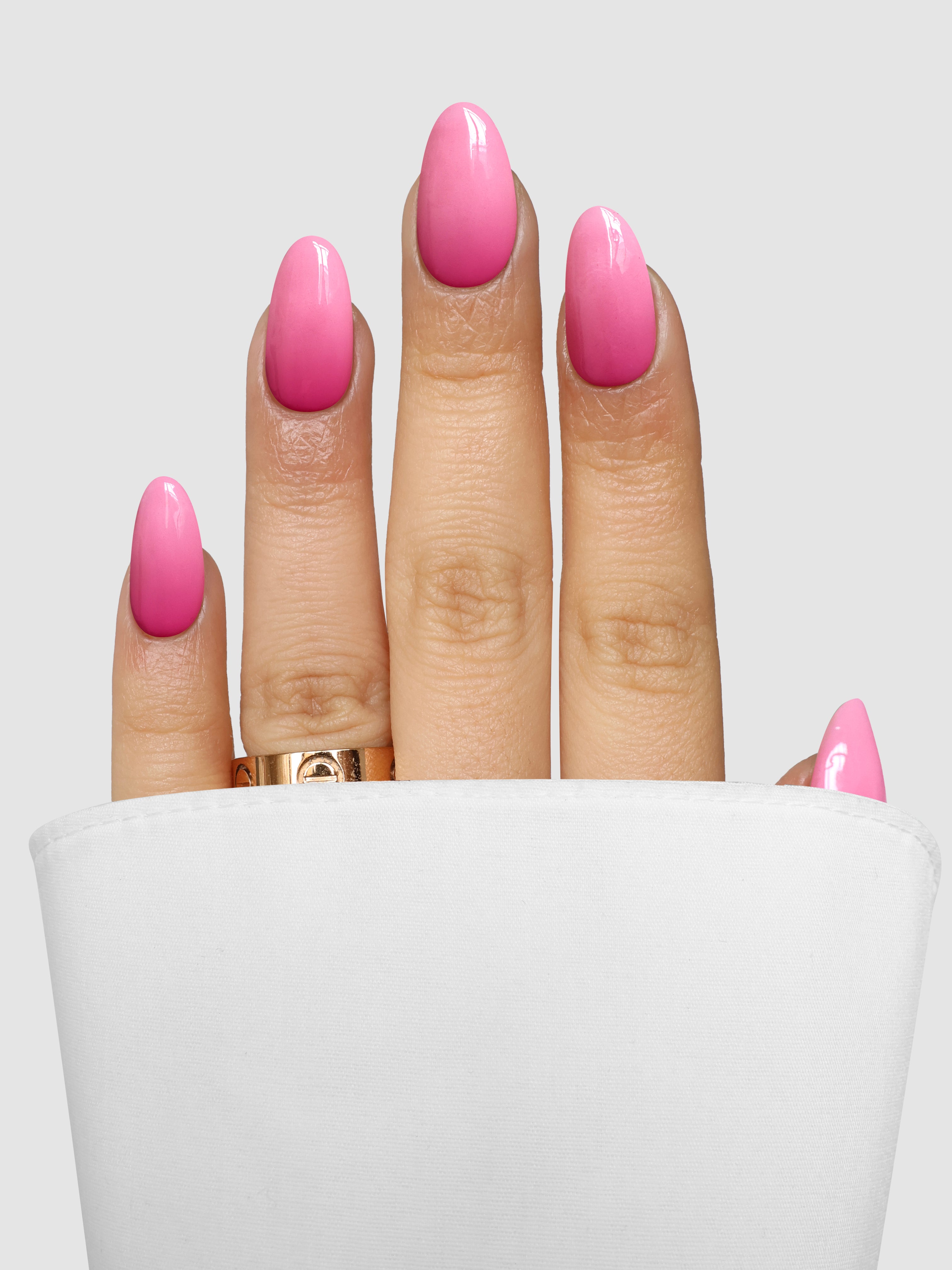 A hand with long, pink ombre nails done using Sassy Saints' Pink Parfait Dipping Powder and adorned with a gold ring is partially covered by a white cloth, set against a light grey background.