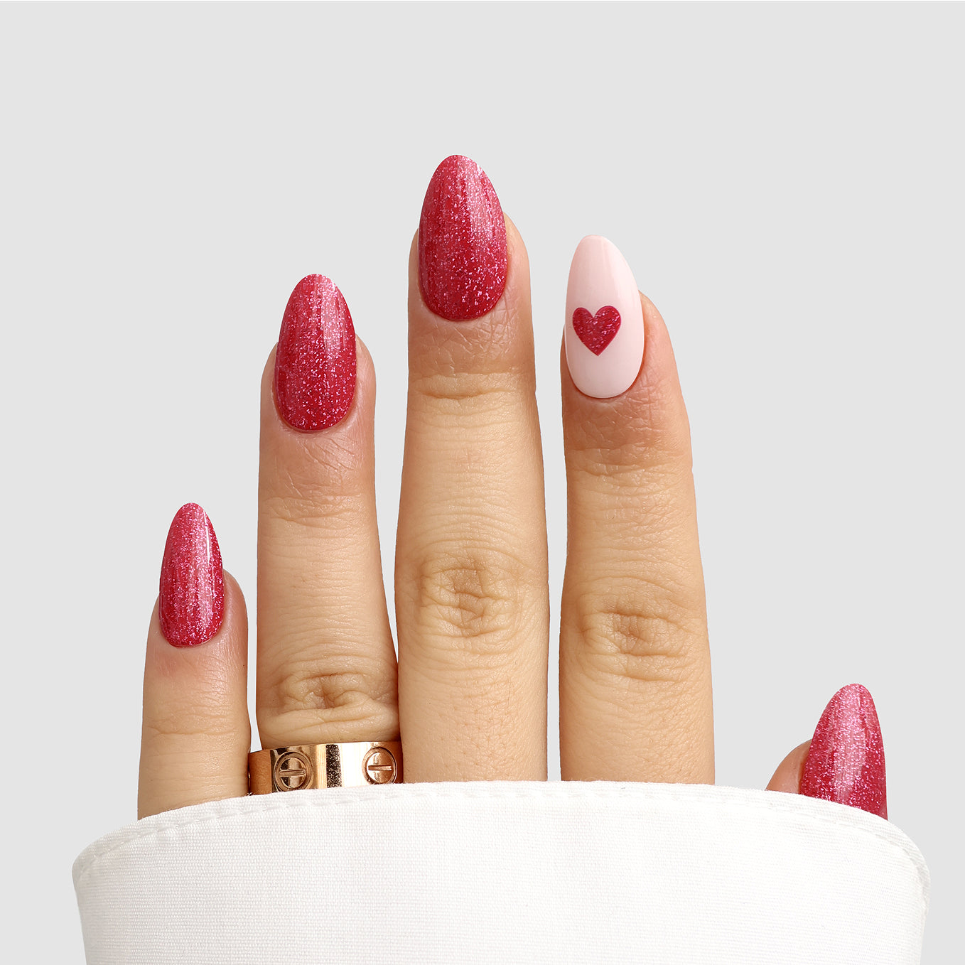 A hand with almond-shaped nails features Sassy Saints' Modern Romance Gel Wrap: four glittery pink nails and a ring fingernail in pale pink with a red heart. The person wears two gold rings and a white sleeve partially covers the hand.