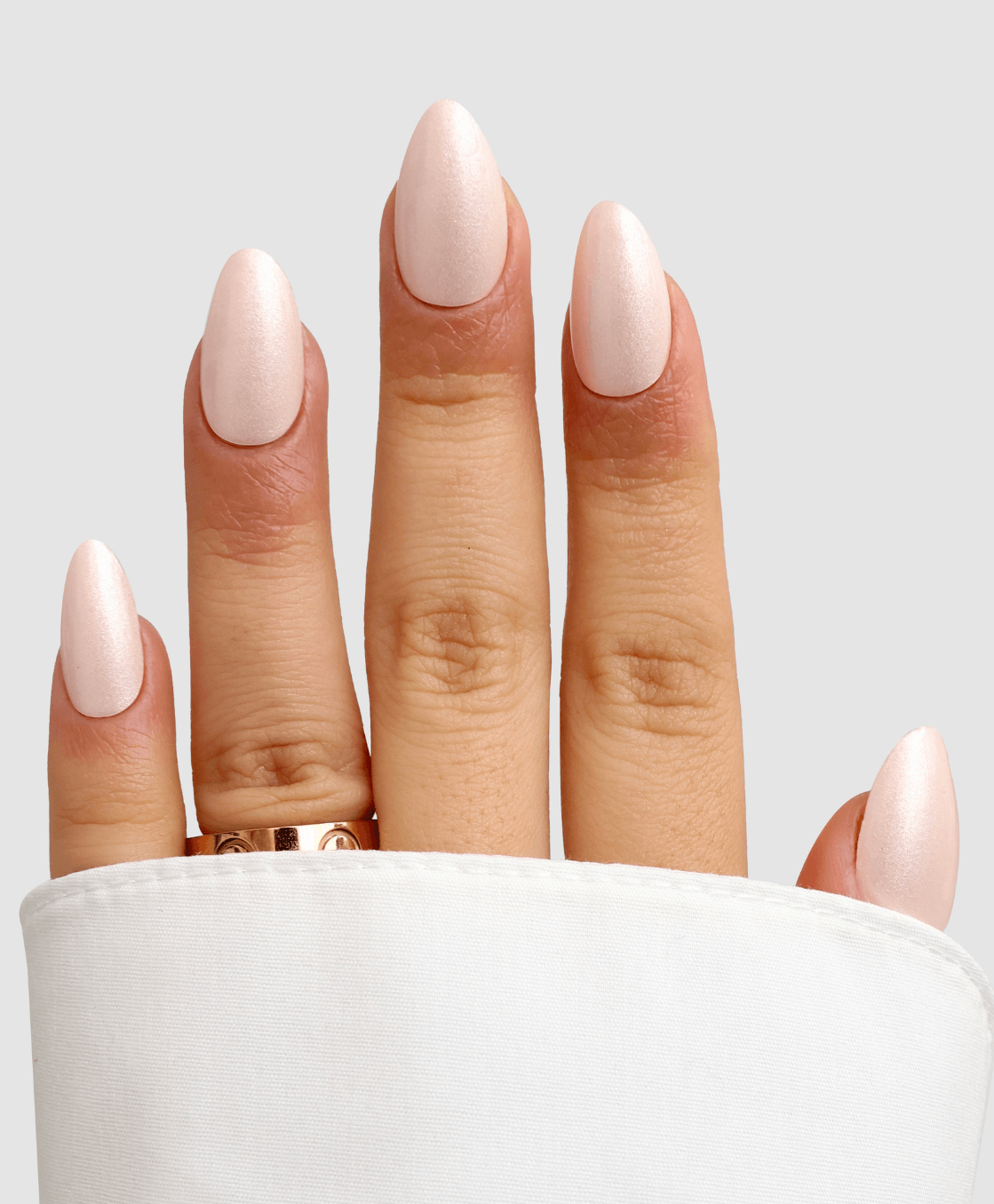 A hand with Sassy Saints Powder Puff Gel Wraps features light pink, almond-shaped nails partially covered by a white sleeve. A gold ring adorns the ring finger against a plain, light gray background. Brings The Salon Home.