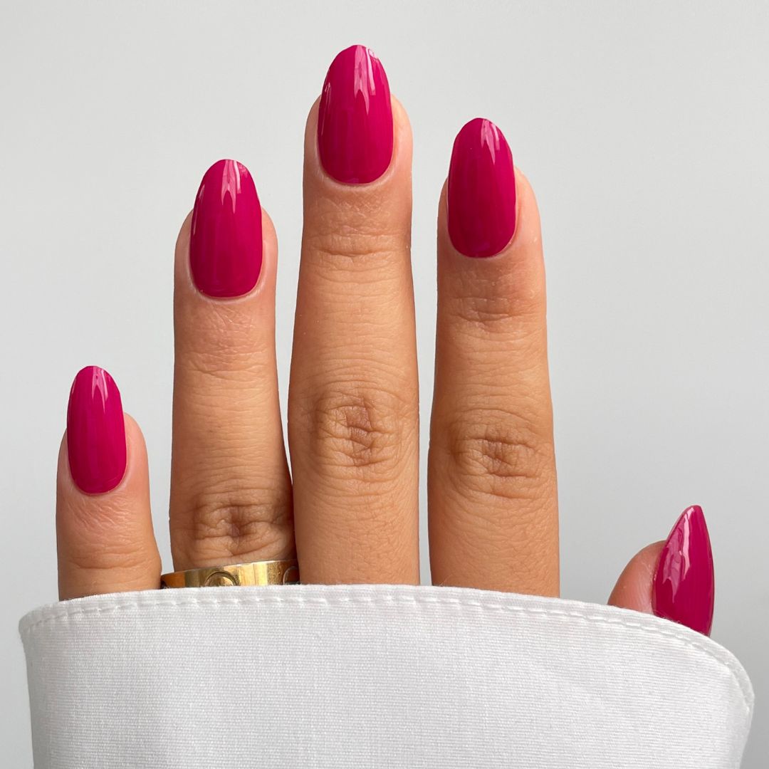 A hand with medium-length, oval nails features Sassy Saints’ Rosewater Silk Gel Wrap in a glossy magenta hue, partially hidden by a white sleeve. A gold ring adorns the middle finger.
