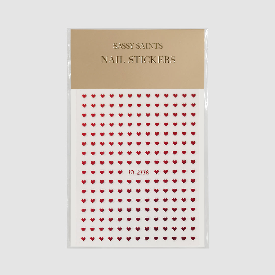 A sheet of Sassy Saints - Brings The Salon Home Red Hearts Nail Stickers with multiple rows of shiny red heart shapes shown in its packaging, displayed on a beige background.