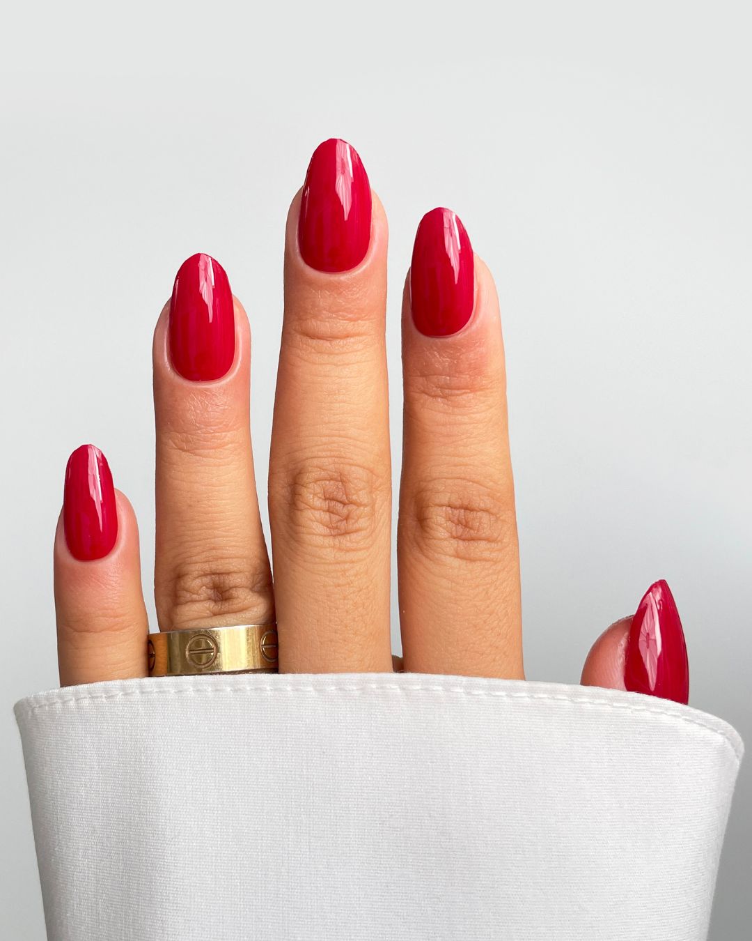 A hand with long, oval nails in Sassy Saints' Red Alert Gel Wrap peeks from a white sleeve, flaunting a wide gold ring on the middle finger against a light gray background. Brings The Salon Home.