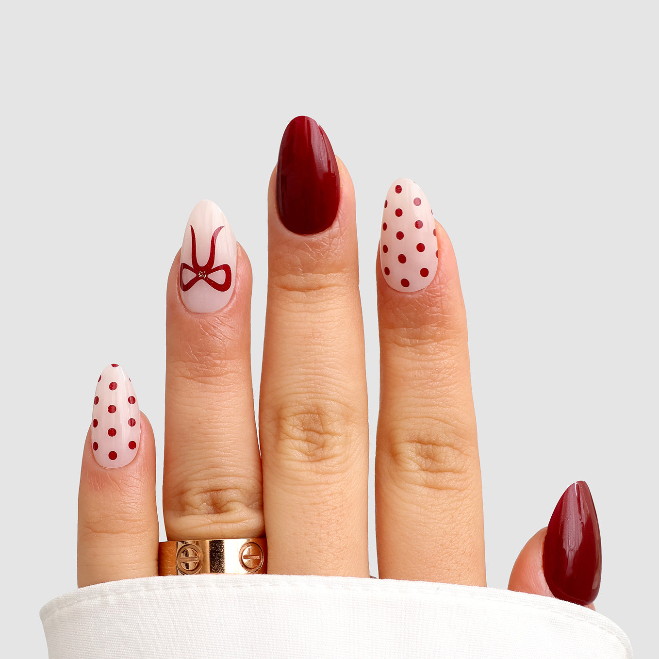 A hand with almond-shaped nails showcases Sassy Saints - Brings The Salon Home's Ribbon Muse Gel Wrap, featuring solid red, red polka dots on white, and a red ribbon accent. Two gold rings and a white sleeve complete the look.