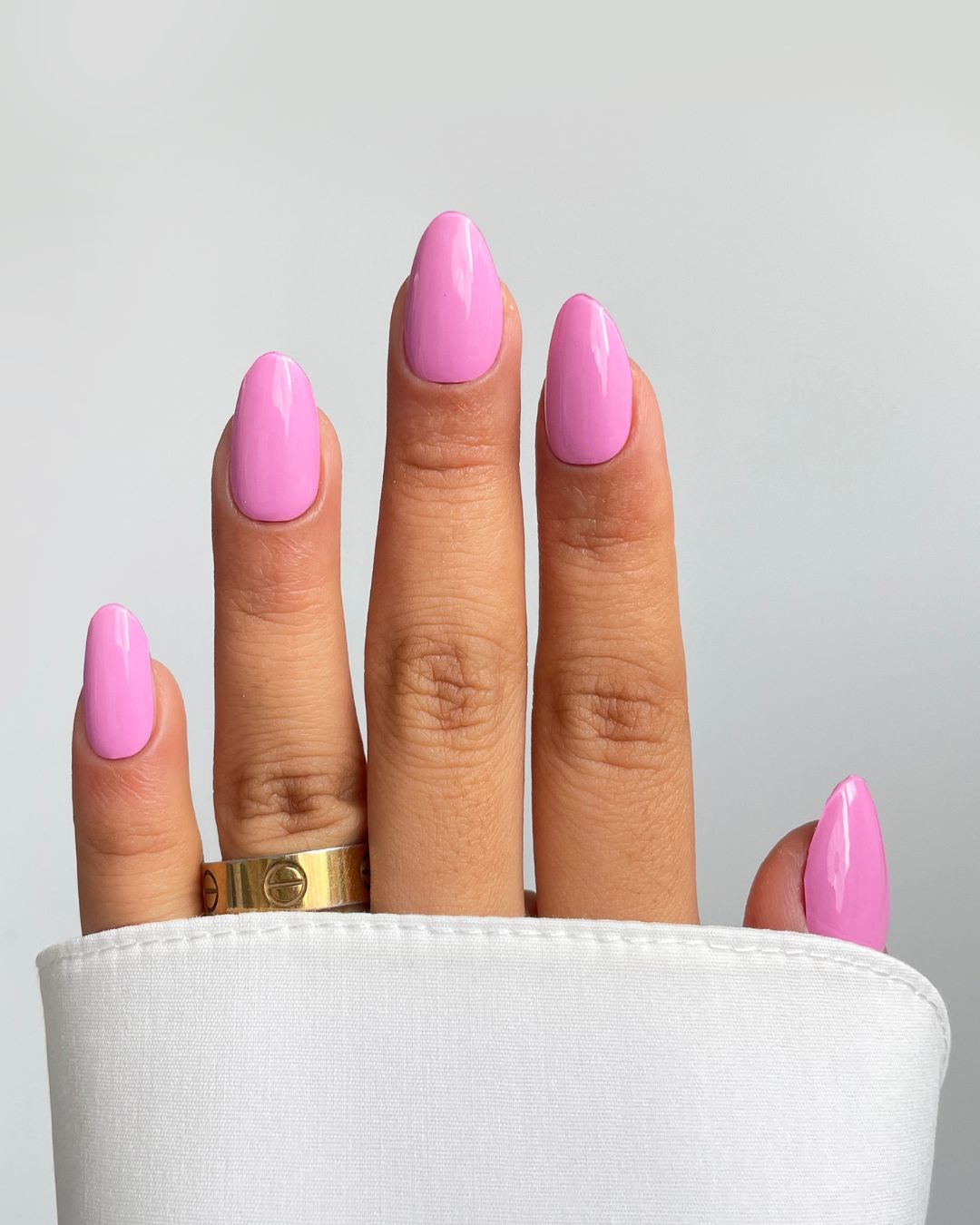 A hand with short, almond-shaped nails features Sassy Saints’ Riviera Pink Gel Wraps—glossy light pink. A gold ring sits on the middle finger, a white sleeve covers the wrist, and the background is plain light gray.