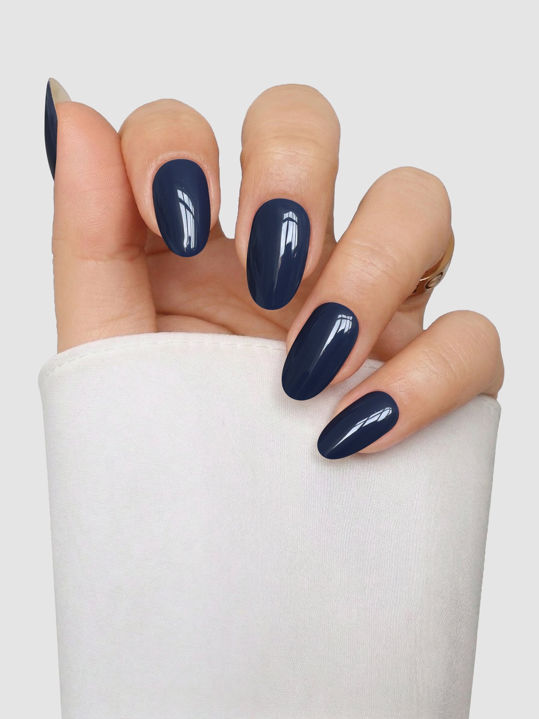 A hand with neatly manicured oval nails features the glossy dark blue “Slate State Gel Wrap” from Sassy Saints – Brings The Salon Home, set against a plain light background and paired with a white long-sleeve garment.