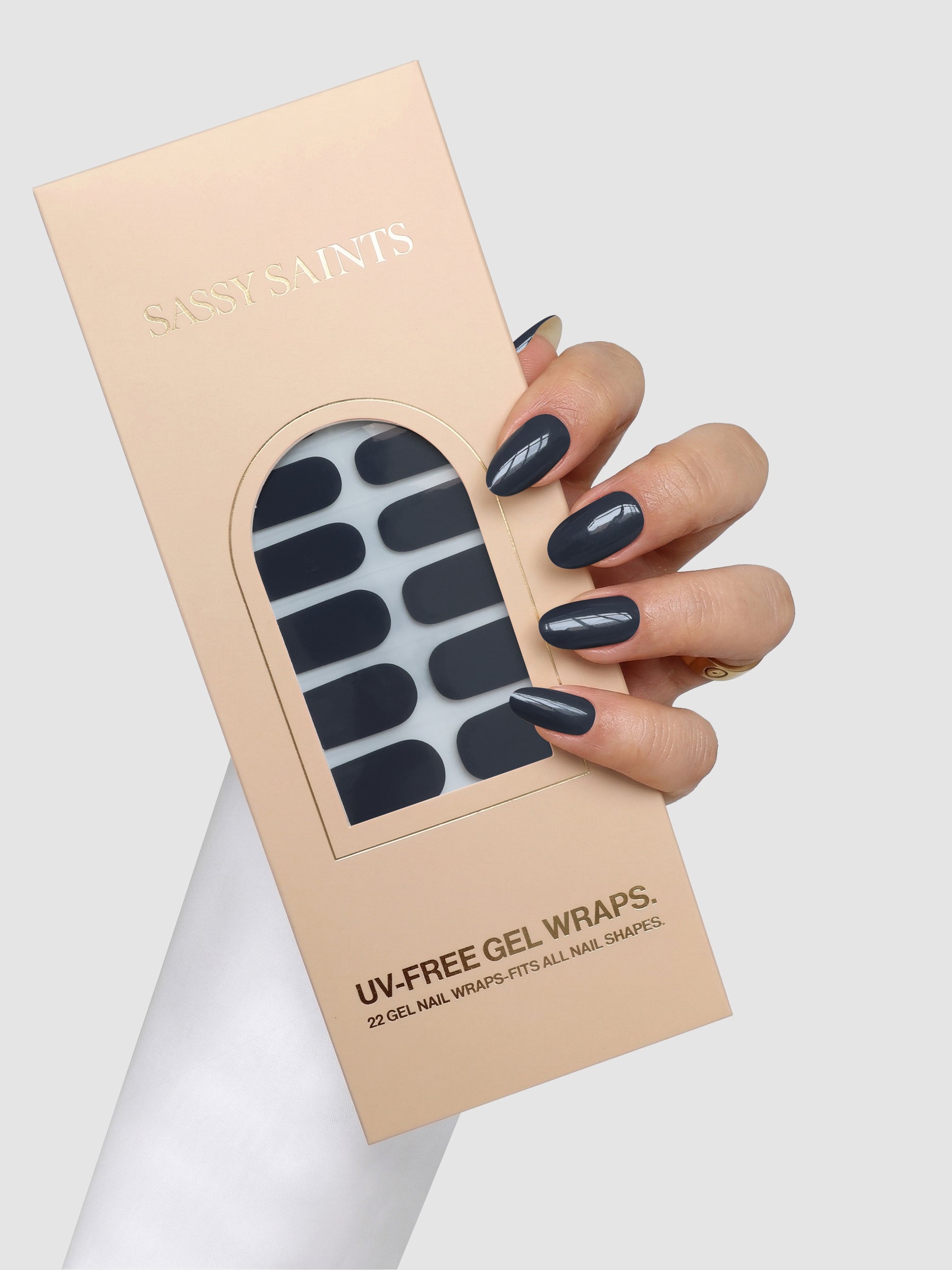 A hand with dark gray polished nails holds a Sassy Saints - Brings The Salon Home Smoke & Mirrors Gel Wrap box, displaying the gel wraps through a clear window on the beige packaging.