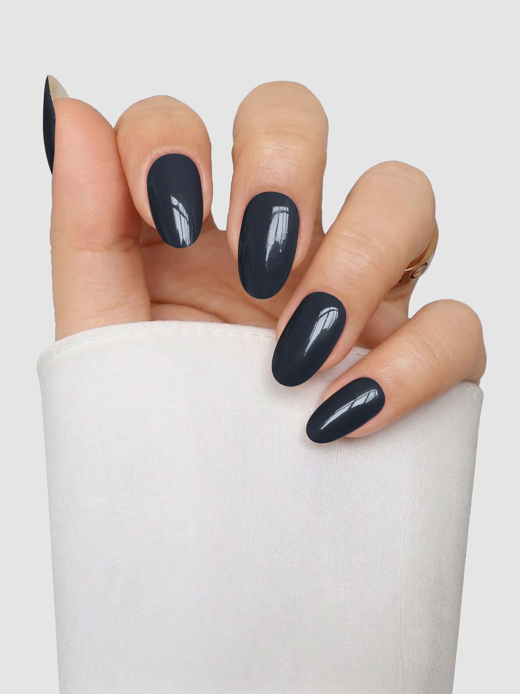 A hand with oval-shaped nails featuring Sassy Saints - Brings The Salon Home’s Smoke & Mirrors Gel Wrap, a glossy dark navy blue finish, is posed against a plain light background. The person is wearing a long-sleeved white top.