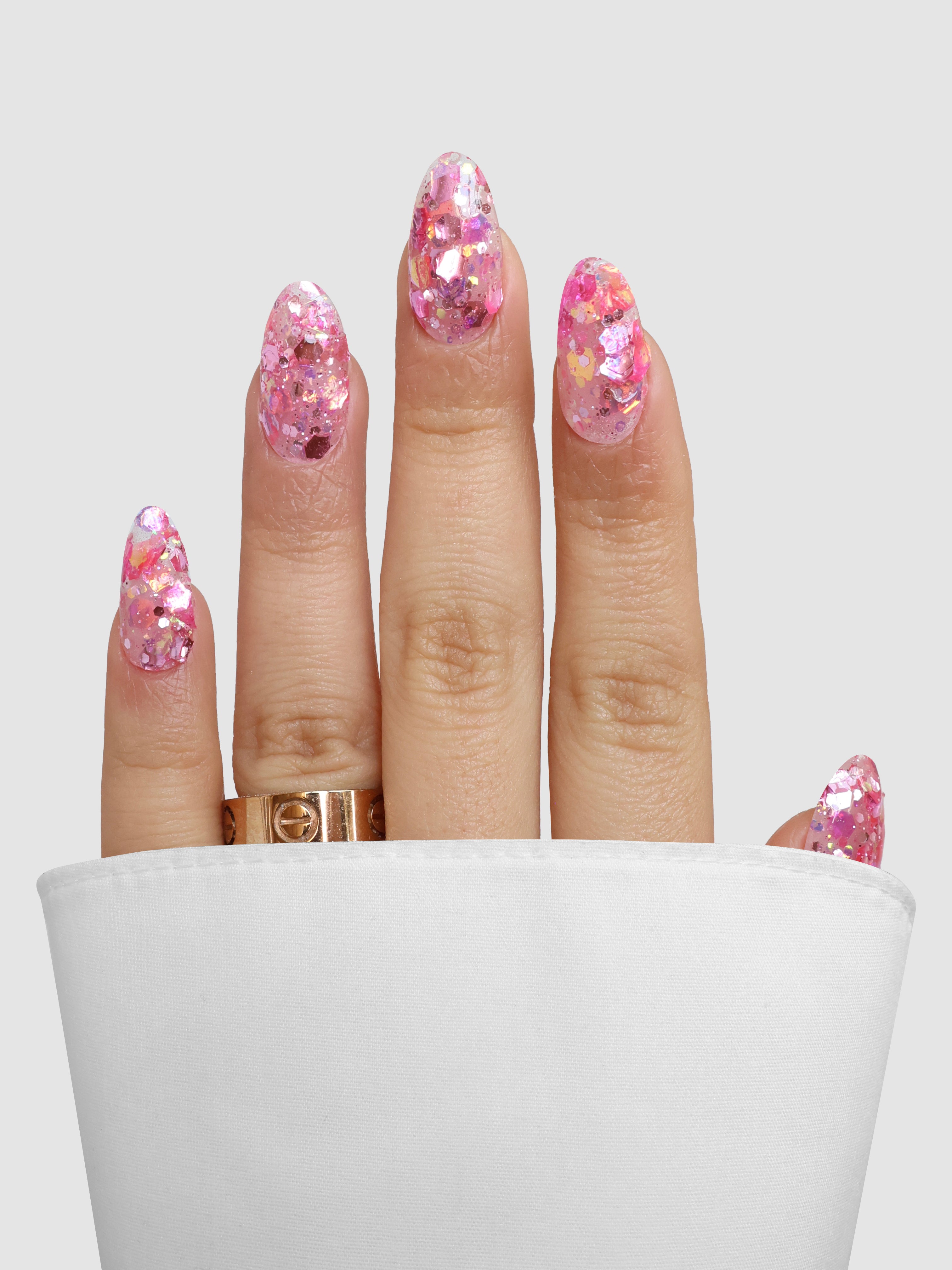 A hand with long, almond-shaped nails in sparkling pink Spring Rose Dipping Powder by Sassy Saints - Brings The Salon Home, wearing a gold ring, is partially tucked into a white shirt cuff against a simple light background.