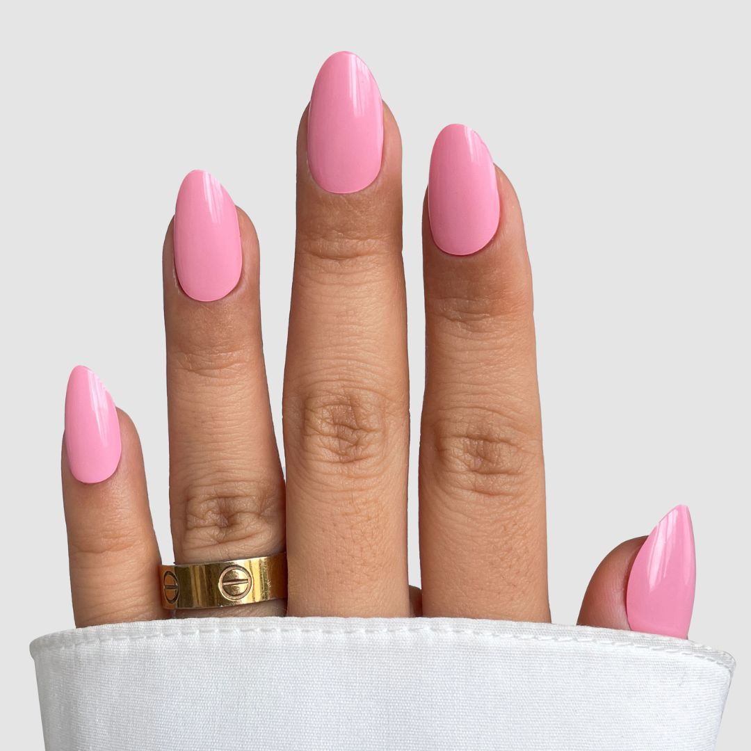 A hand with long, almond-shaped nails features Sassy Saints' Sugar Candy Gel Wrap glossy pink, paired with a gold ring and partially covered by a white sleeve against a plain light gray background. Brings The Salon Home.