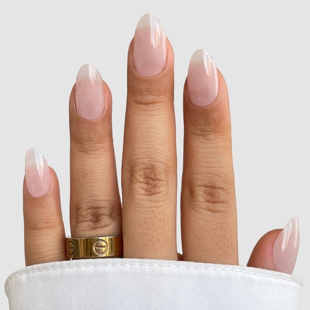 A hand shows off long, almond-shaped nails in Sugar Glaze Gel Wrap by Sassy Saints - Brings The Salon Home, featuring a natural pink base with white tips. A gold ring adorns the middle finger, and a white sleeve covers part of the wrist.