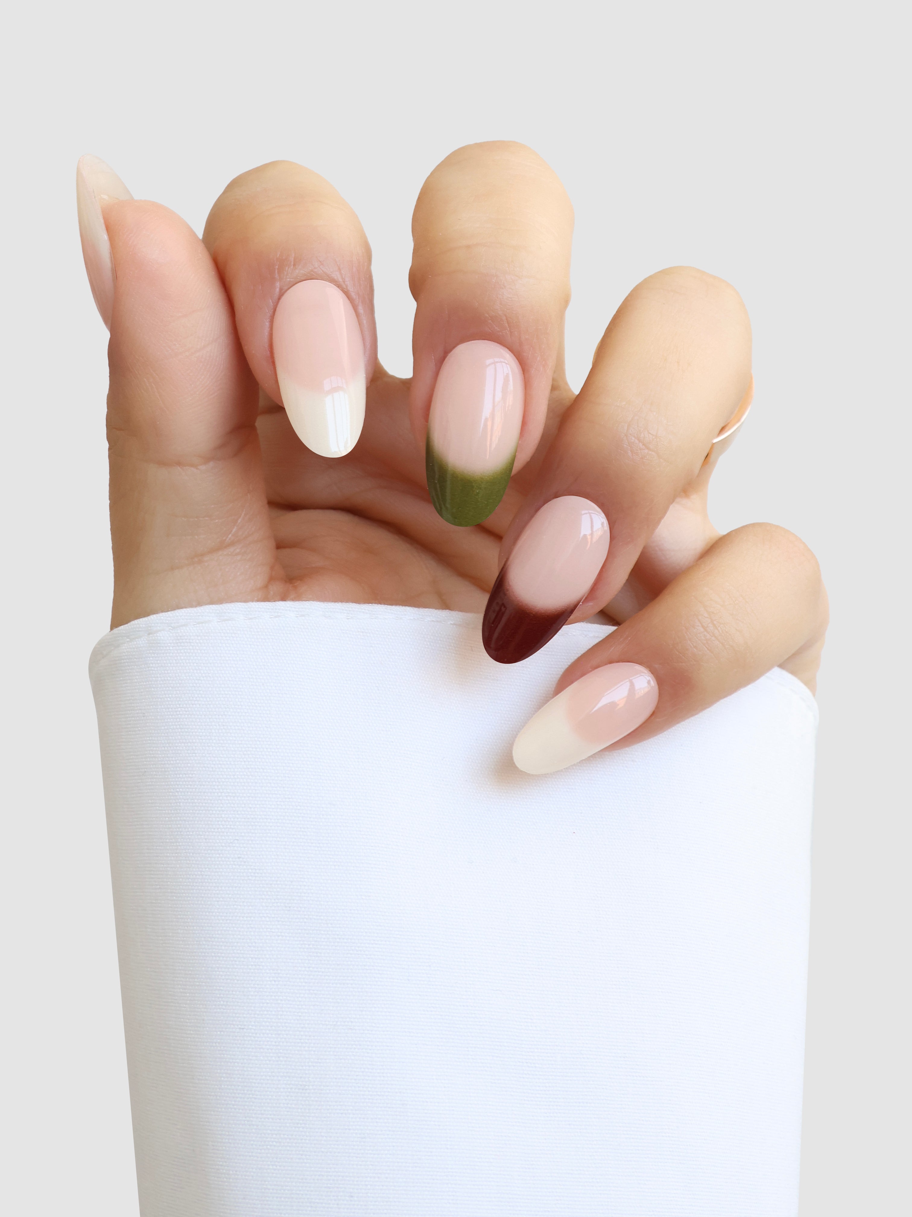 A hand with long, almond-shaped nails shows off Sassy Saints' Terre Chic Gel Wraps: three French tips in white, one tip in green, and one in dark maroon. The look is completed by a white sleeve partially covering the hand.