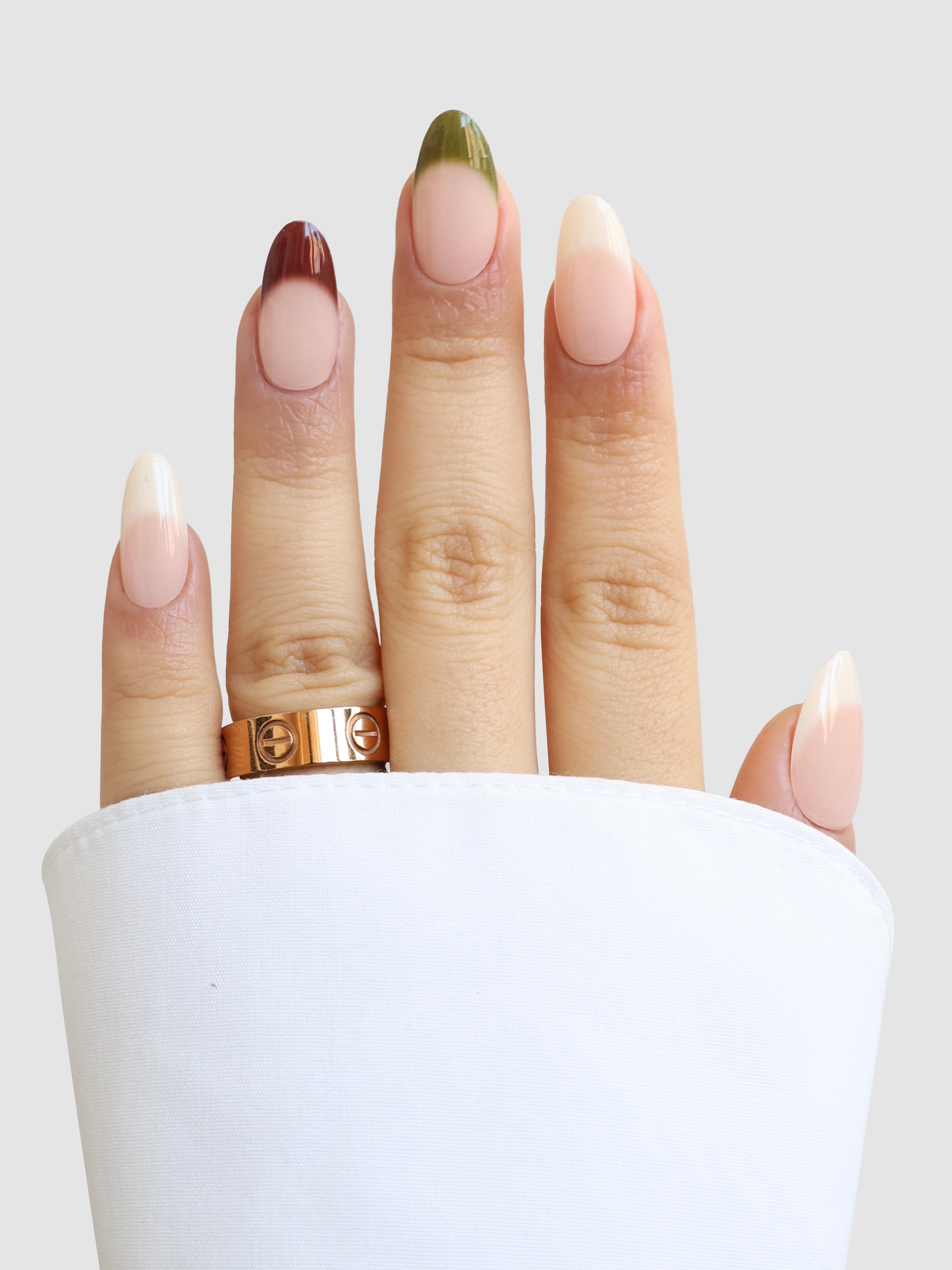 A hand with long, almond-shaped nails showcases Sassy Saints' Terre Chic Gel Wraps—each tip a unique earth tone. Two gold rings and a white cuffed sleeve complete the minimalist, salon-quality look at home.