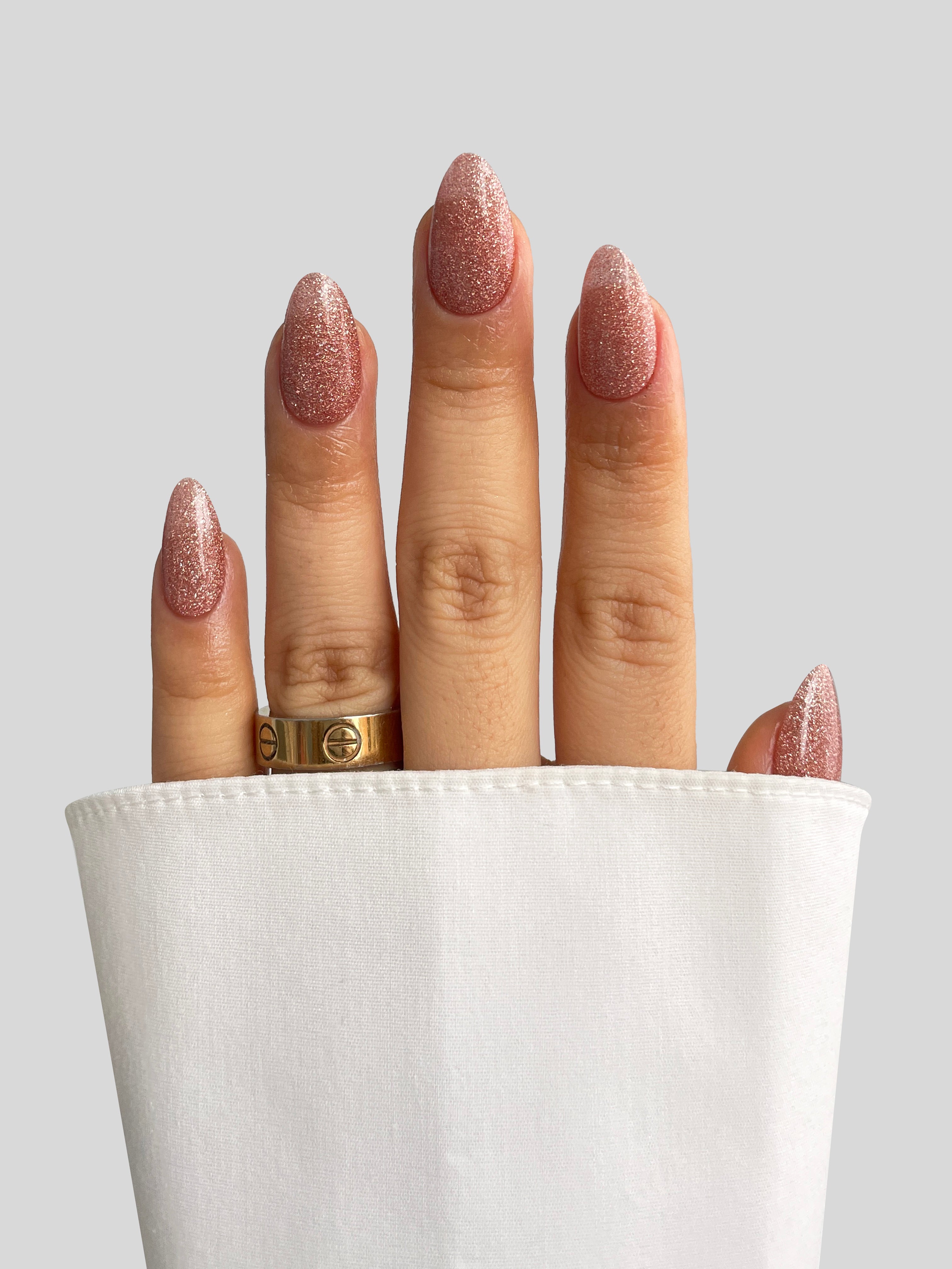 A hand with long, glittery pink nails created using Sassy Saints - Brings The Salon Home’s Toasted Tinsel Dipping Powder and a gold ring is partially covered by a white sleeve against a plain light gray background.