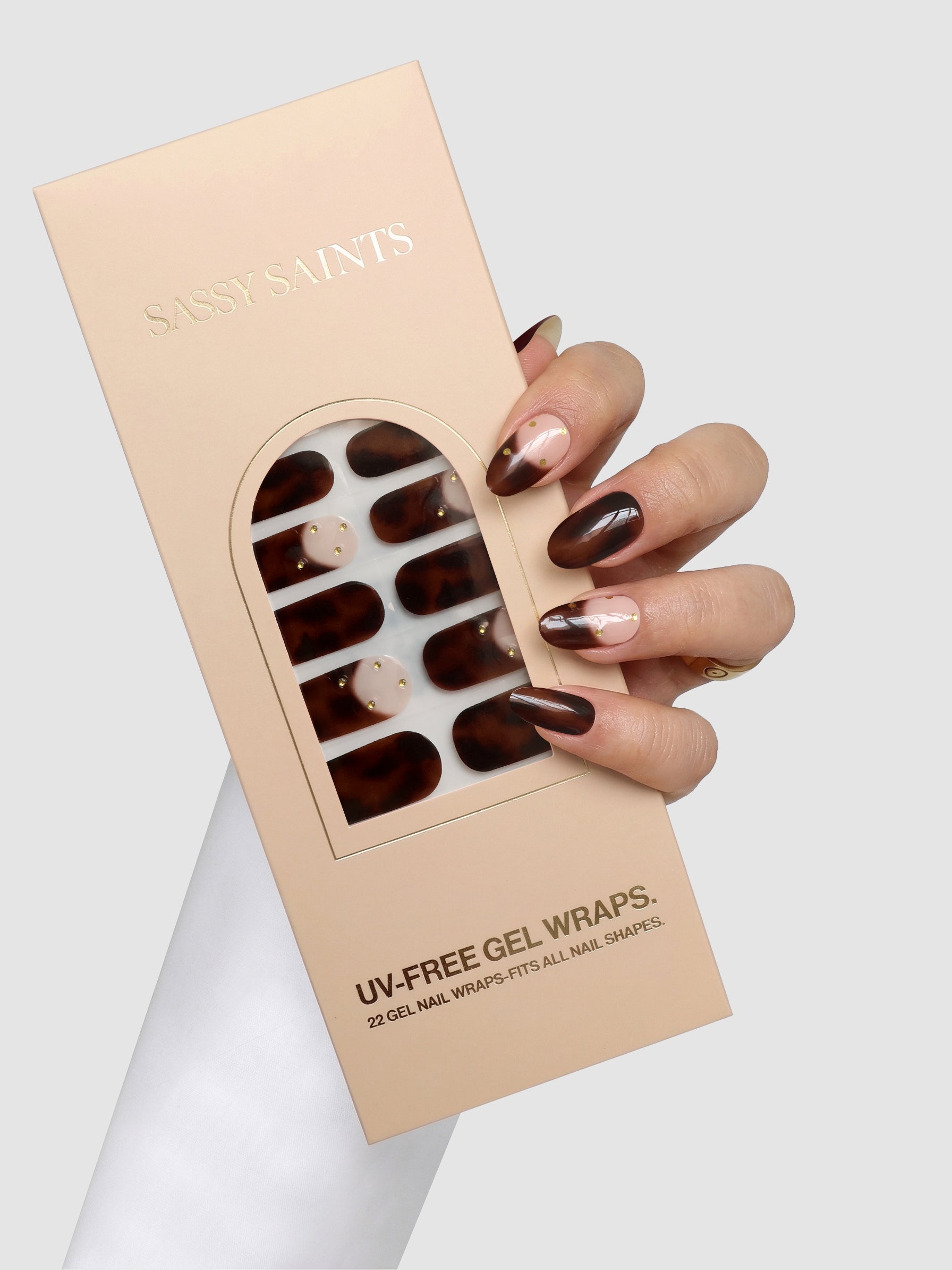 A hand with brown and pink nail art holds a box of Sassy Saints - Brings The Salon Home Tortoise Takeover Gel Wraps in brown and nude shades against a plain light background.