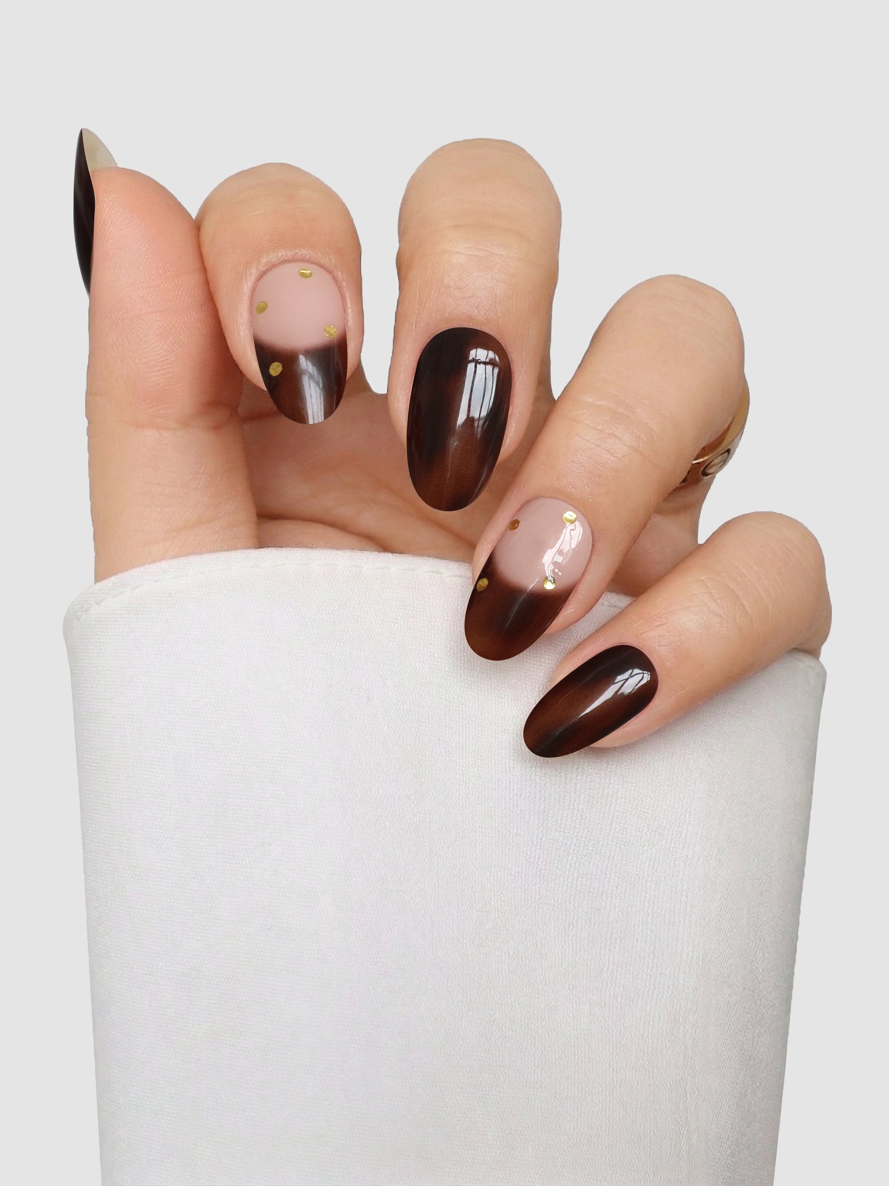 A hand with almond-shaped nails styled using Sassy Saints - Brings The Salon Home’s Tortoise Takeover Gel Wrap, featuring a brown gradient French tip design accented with gold studs and subtle line art, posed against a light gray background. The wrist is partially covered by a white sleeve.