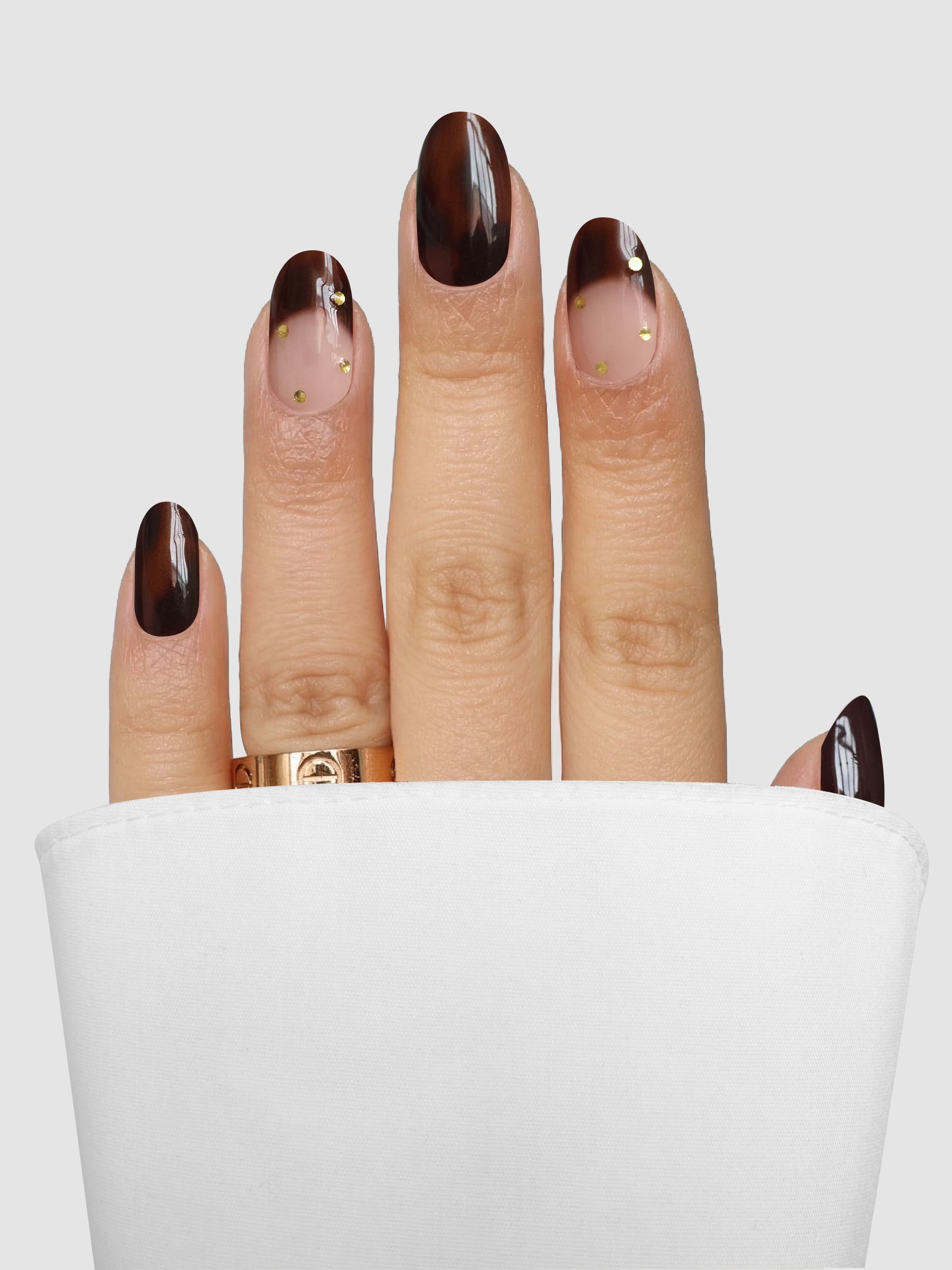A hand adorned with Sassy Saints - Brings The Salon Home’s Tortoise Takeover Gel Wrap, featuring a brown gradient design and small gold dots, rests against a white cloth. The wearer sports a gold ring on the middle finger, set against a light gray background.