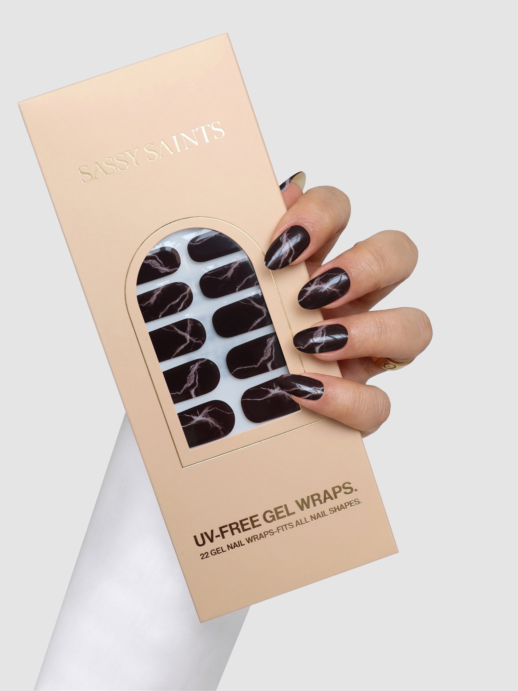 A hand with black marble-patterned Truffle Swirl Gel Wraps holds a beige Sassy Saints – Brings The Salon Home box labeled UV-Free Gel Wraps, containing matching nail wraps inside.