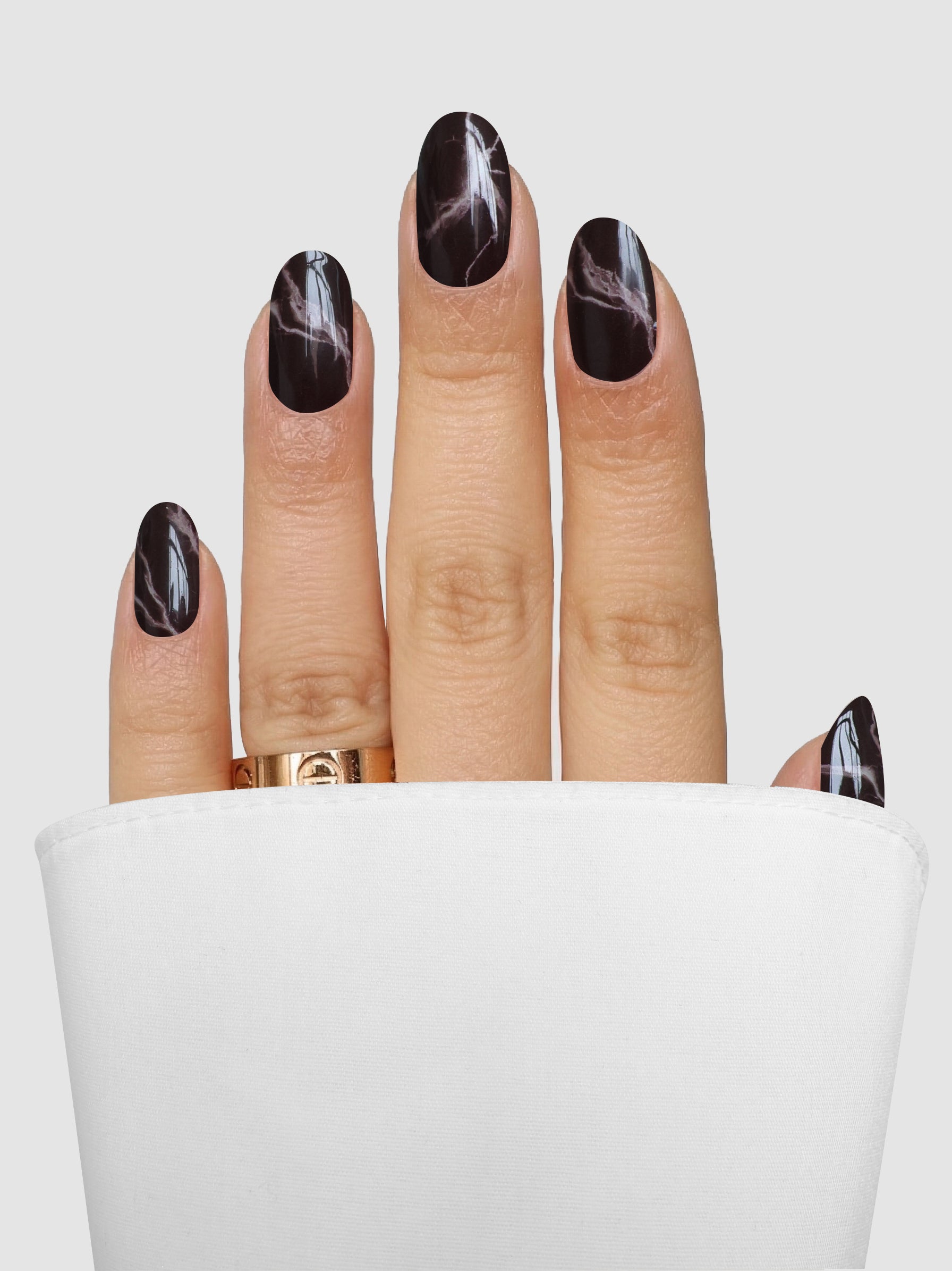 A hand adorned with Sassy Saints - Brings The Salon Home’s Truffle Swirl Gel Wrap features dark, glossy, marble-patterned nails and a gold ring, partially tucked into a white cuff against a light gray background.