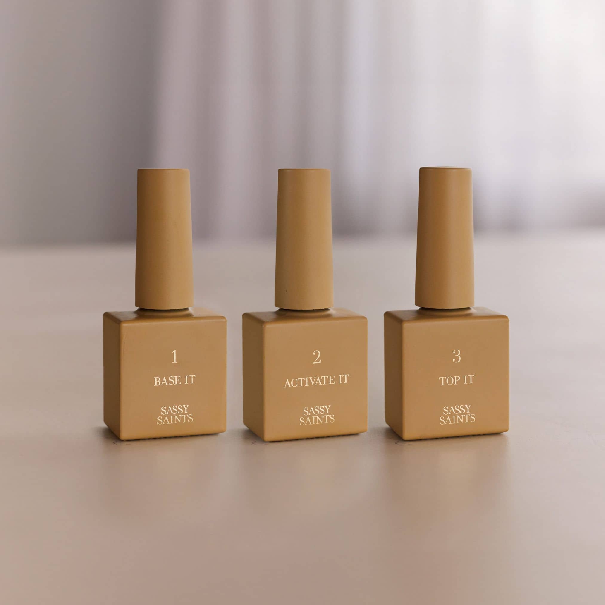 Three beige bottles from Sassy Saints - Brings The Salon Home, labeled 1 BASE IT, 2 ACTIVATE IT, and 3 TOP IT, are lined up on a light surface. This is the Refill Bundle (NEW), shown with a blurred background.