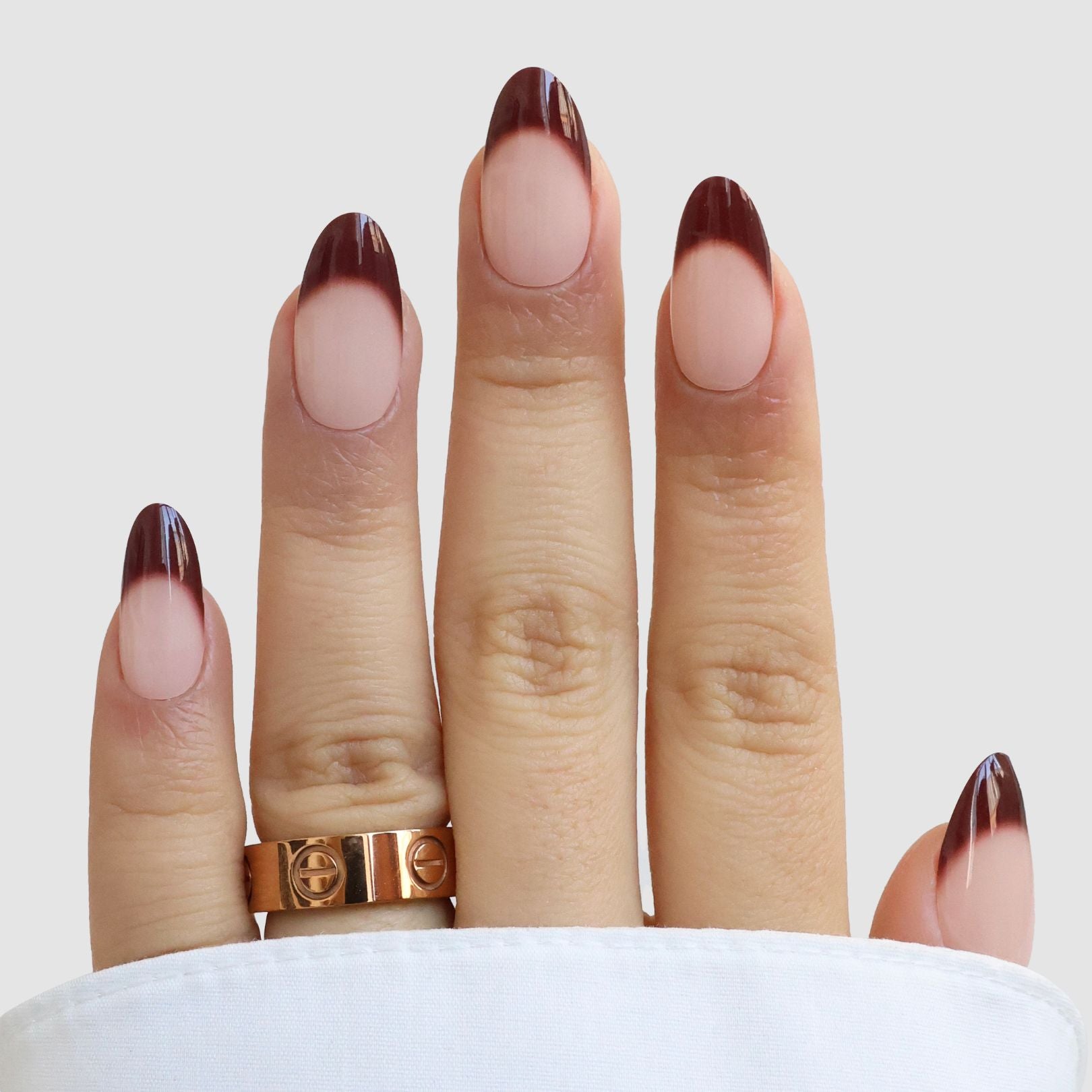 A hand with almond-shaped nails wears Sassy Saints’ Haute Bordeaux Gel Wraps, featuring a nude base and dark brown French tips. Two gold rings and a white sleeve are visible against a plain light gray background.
