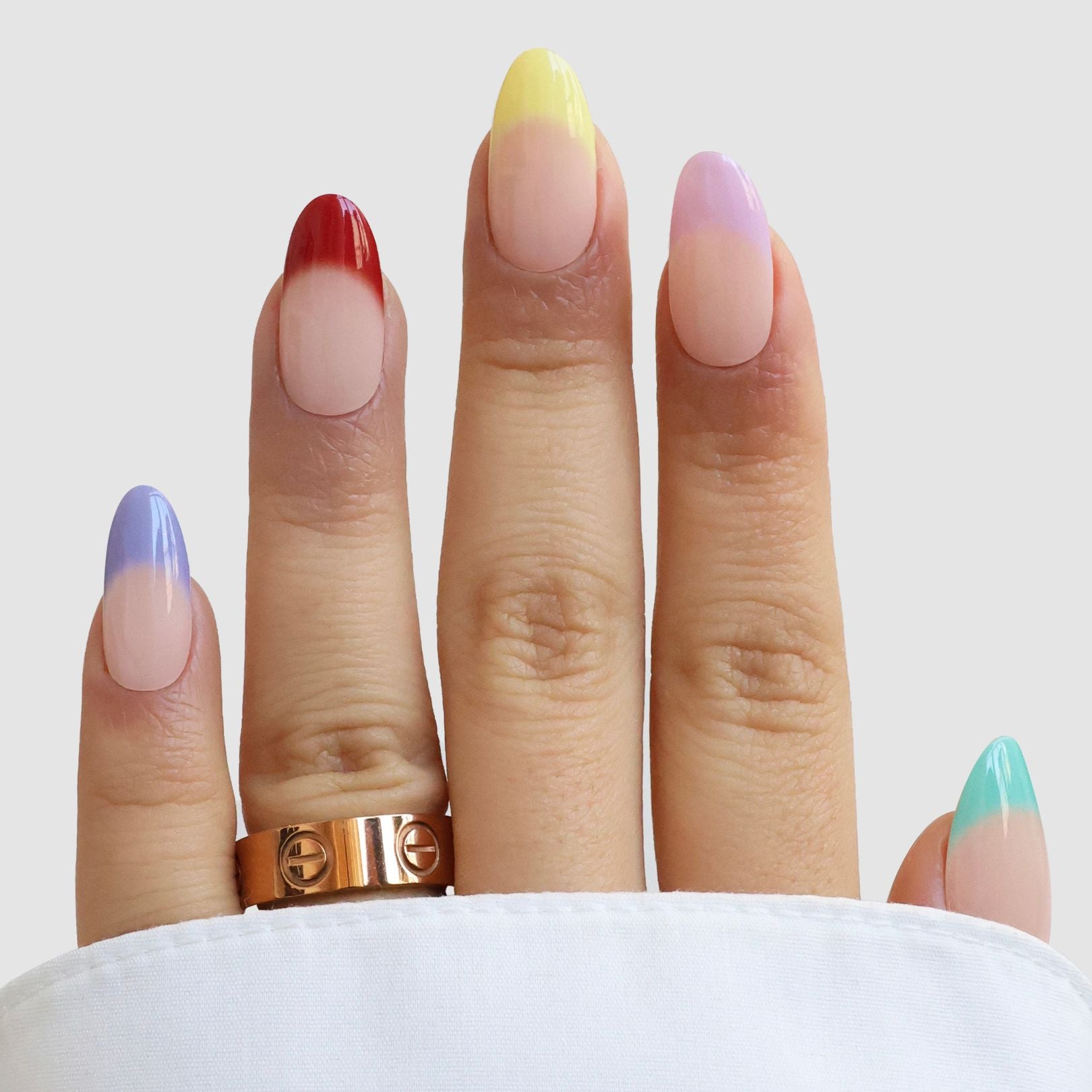 A hand with almond-shaped nails displays Sassy Saints - Brings The Salon Home’s Jardin de Couleurs Gel Wraps, each nail featuring a unique pastel gradient tip in red, purple, yellow, pink, and teal. A gold ring and white sleeve complete the look.