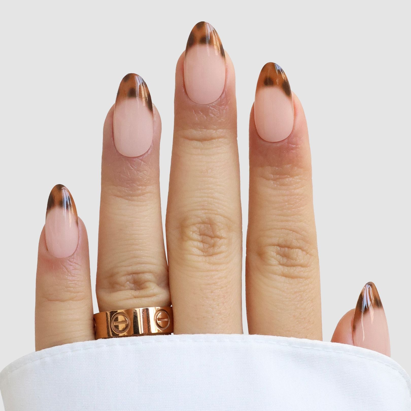 A hand shows off almond-shaped nails with Écaille de Luxe Gel Wrap by Sassy Saints—nude base and tortoiseshell tips—plus two gold rings and a white cuffed sleeve.