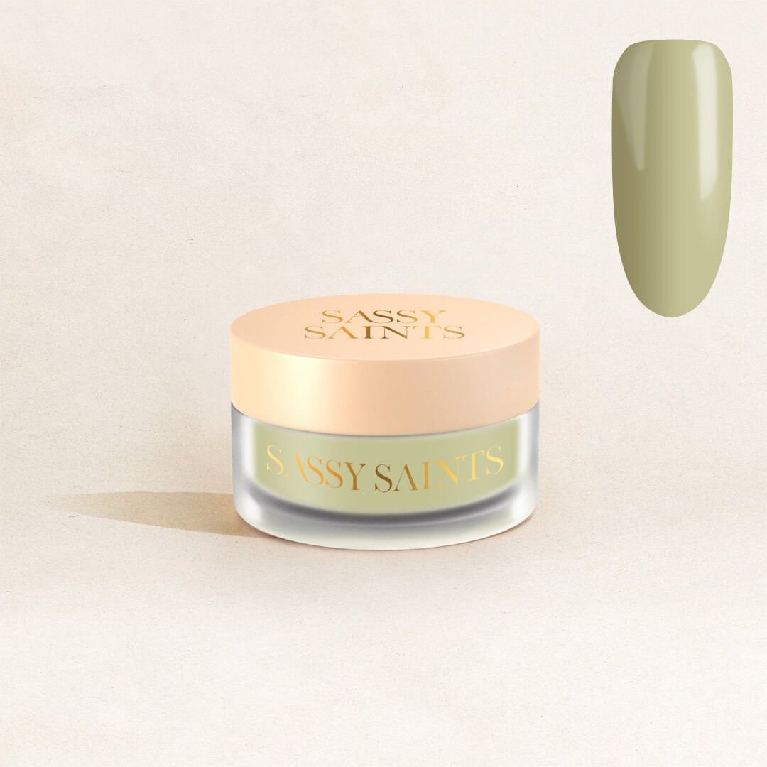A small frosted glass jar with a beige lid labeled "Sassy Saints - Brings The Salon Home" sits on a light surface. To the right, a swatch of olive green Muted Moss Dipping Powder is displayed.
