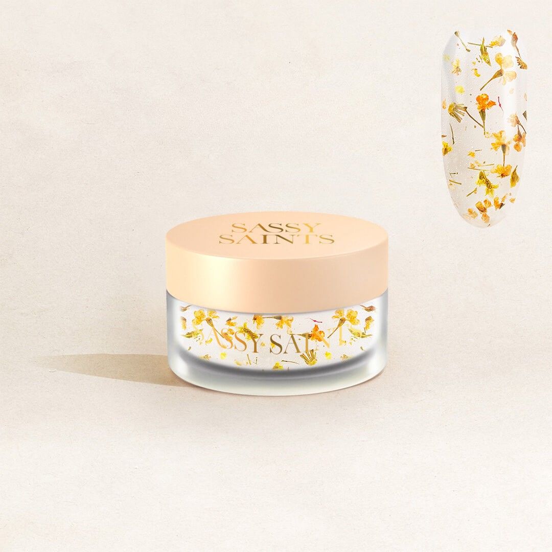 A frosted glass jar labeled Sassy Saints - Brings The Salon Home with a beige lid and yellow-orange dried flowers sits on a light background; above it, a nail swatch features Marigold Mirage Dipping Powder in matching floral designs.