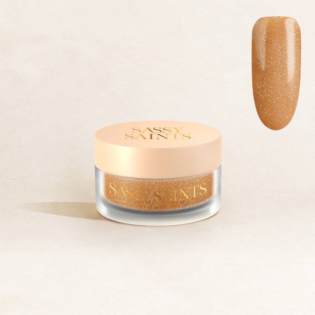 A small round jar labeled Sassy Saints - Brings The Salon Home holds Golden Goddess Dipping Powder, a shimmery gold powder. Next to it, a close-up swatch displays its glittery gold effect as seen on a nail.