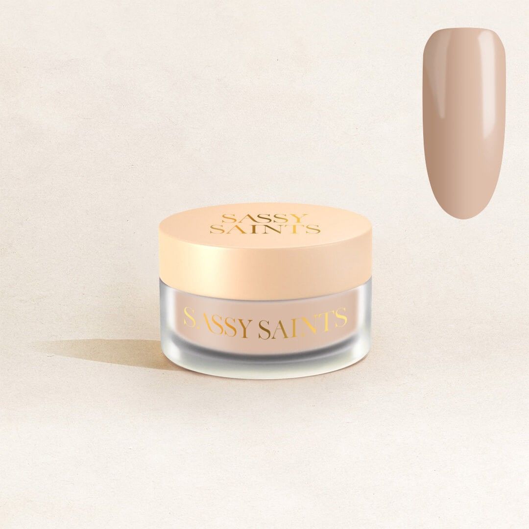 A small round jar with a beige lid labeled Sassy Saints - Brings The Salon Home, featuring Bridal Biscuit Dipping Powder, sits on a light surface. A sample of its glossy, nude beige shade appears in the upper right corner.