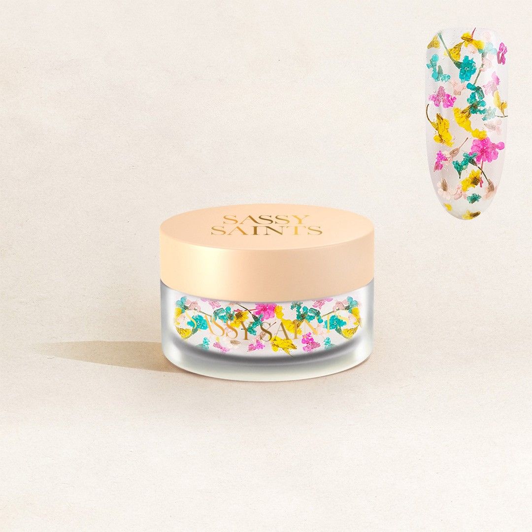 A small frosted jar with a beige lid labeled "Spring Symphony Dipping Powder" by Sassy Saints - Brings The Salon Home sits against a soft beige background, with vibrant floral nail art displayed on a sample beside it.