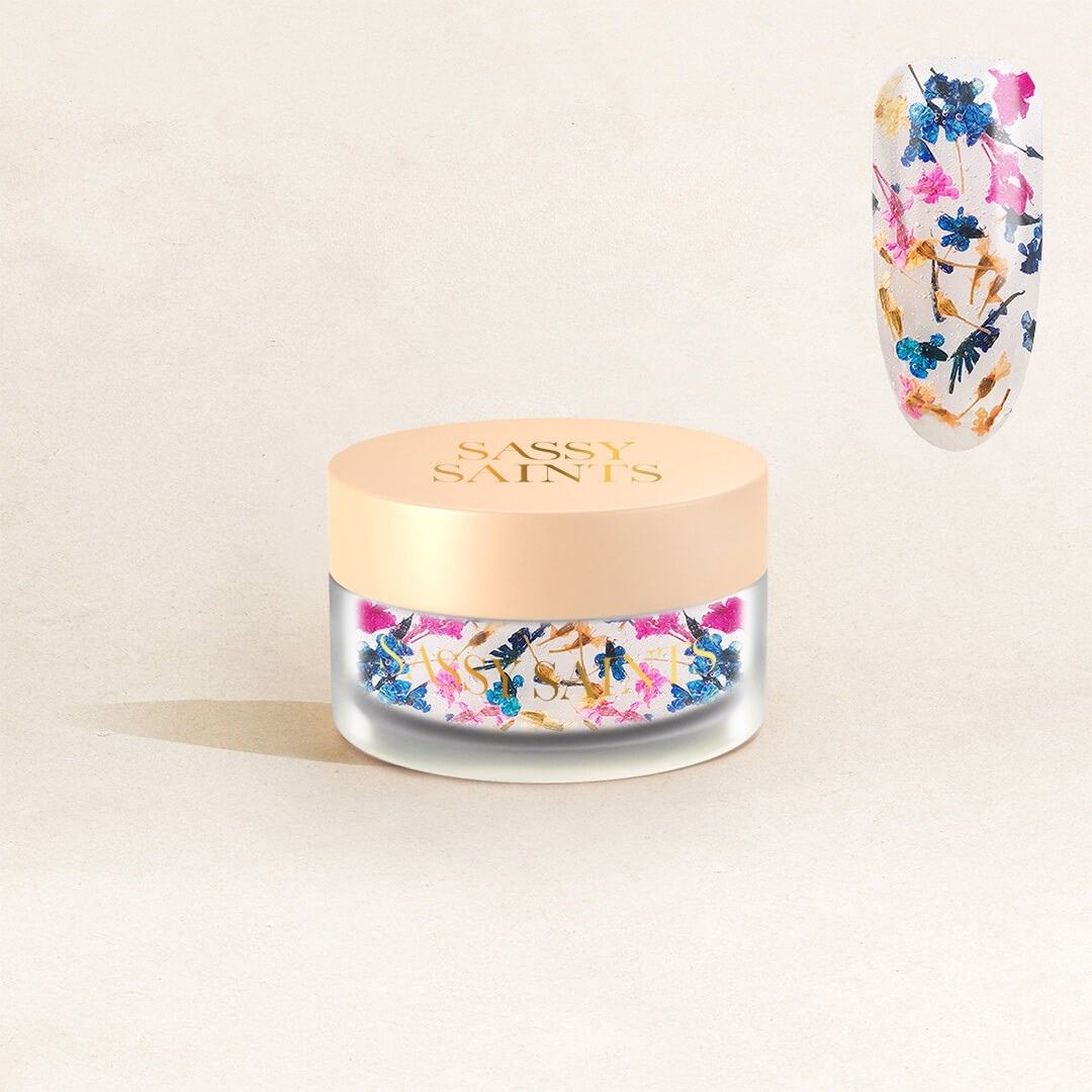 A small frosted jar with a peach lid labeled Sassy Saints - Brings The Salon Home contains Celestial Cascade Dipping Powder. Above, a nail displays vibrant blue, pink, and gold designs made with this powder.