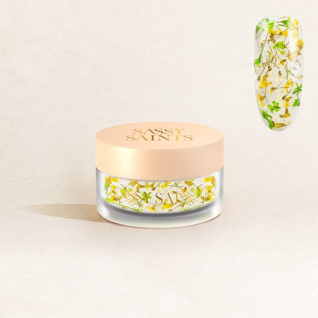 The Wild Whispers Dipping Powder by Sassy Saints - Brings The Salon Home comes in a small jar with a beige lid and features clear powder with yellow flower and green leaf pieces, shown on a nail swatch above.
