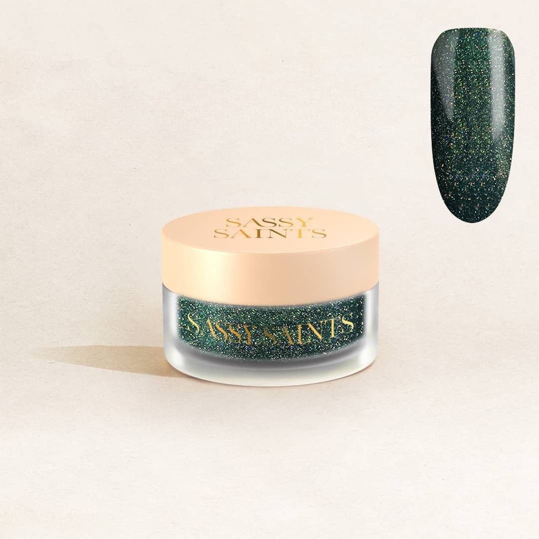 A small jar of Sassy Saints - Brings The Salon Home Emerald Enchantress Dipping Powder with a beige lid sits on a light background. Above, a swatch displays the sparkling dark green powder’s glossy finish.