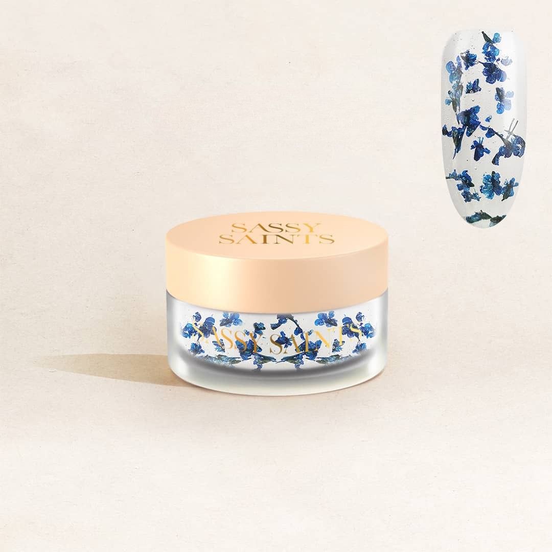 A jar labeled "Sapphire Serenade Dipping Powder" by Sassy Saints - Brings The Salon Home, with a beige lid, sits beside a close-up nail showing the blue powder artfully applied for a decorative manicure.
