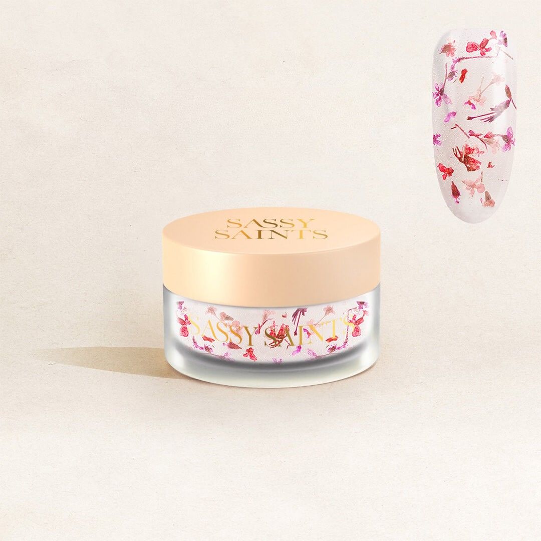 A jar of Rosy Reverie Dipping Powder by Sassy Saints - Brings The Salon Home sits on a light background, with a swatch of clear nail gel adorned with pink and red floral designs shown above and to the right of the jar.