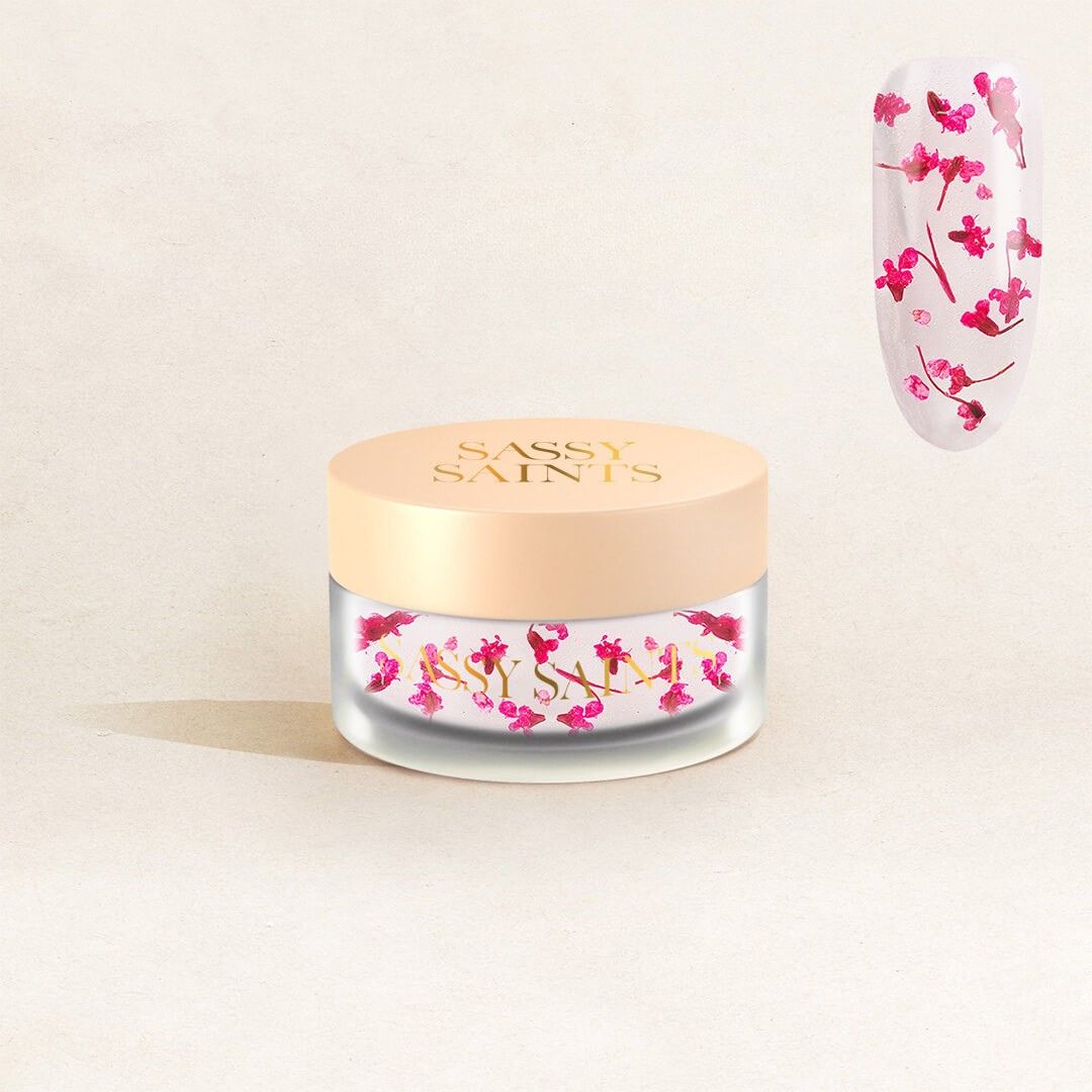 A small jar labeled “Blossom Bliss Dipping Powder” by Sassy Saints – Brings The Salon Home holds pink floral nail art powder, shown beside a close-up of a nail decorated with the same pink blossoms.
