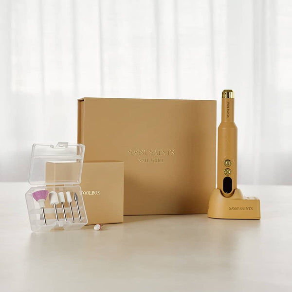 The Sassy Saints At-Home E-File kit includes a gold manicure tool with charger, beige storage box, and small case of drill bits—perfect for salon-quality nail care at home and elegantly displayed on a light background.